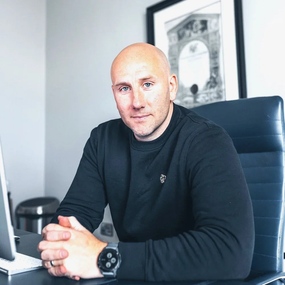 A man with a shaved head sitting at a desk in an office, wearing a black long sleeve shirt and a smartwatch, looking at the camera.