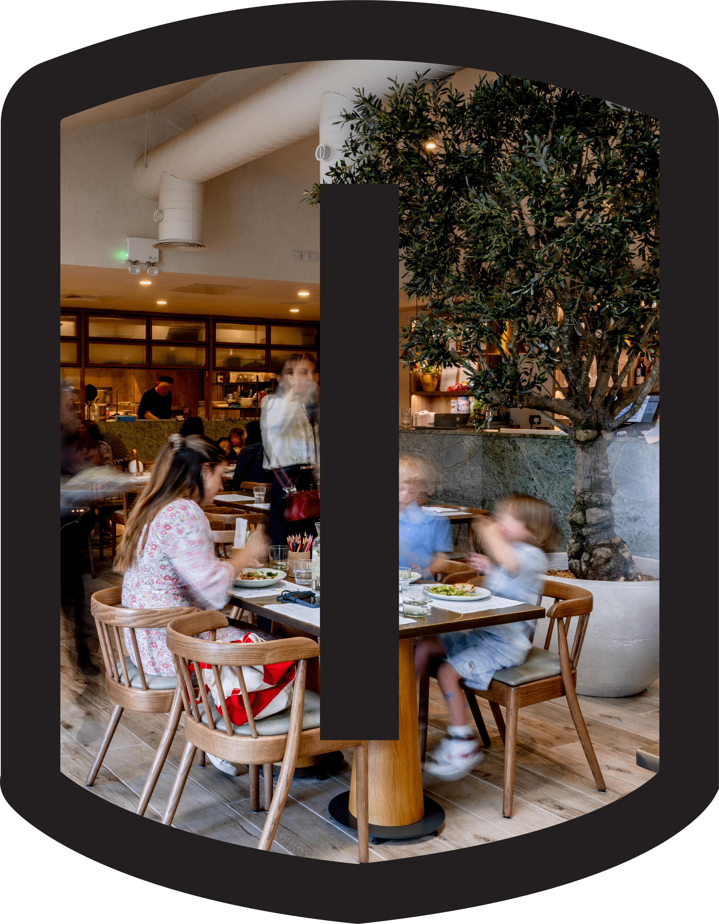 Interior of a restaurant with wooden chairs and tables, a large potted tree, diners dining, and a counter with a kitchen in the background.