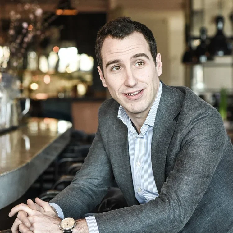 A man with short dark hair and light skin, dressed in a gray suit and a light blue collared shirt, is sitting at a counter in a bar or café, smiling and looking slightly to his right. The background is blurred with warm lighting and bar equipment.
