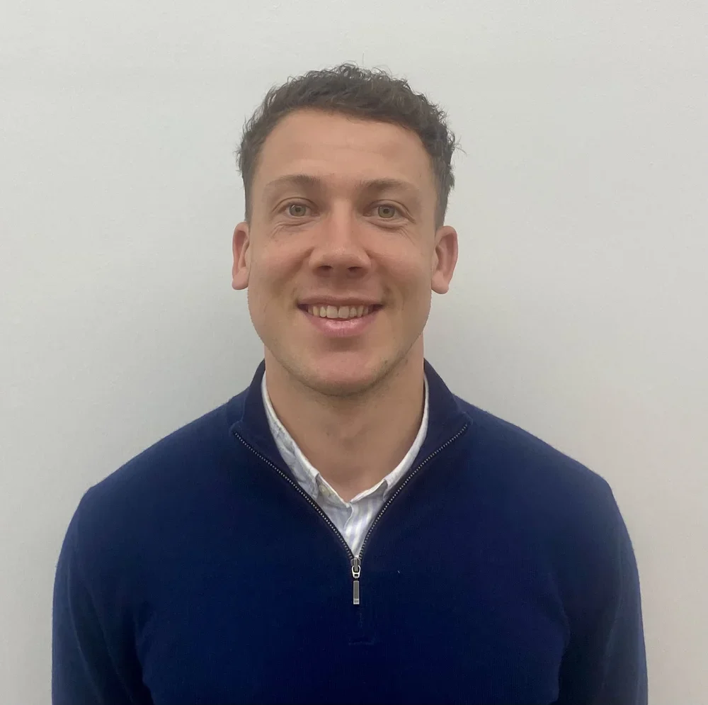 A smiling man with short curly dark hair, wearing a black zip-up sweater over a white collared shirt, standing against a plain white wall.