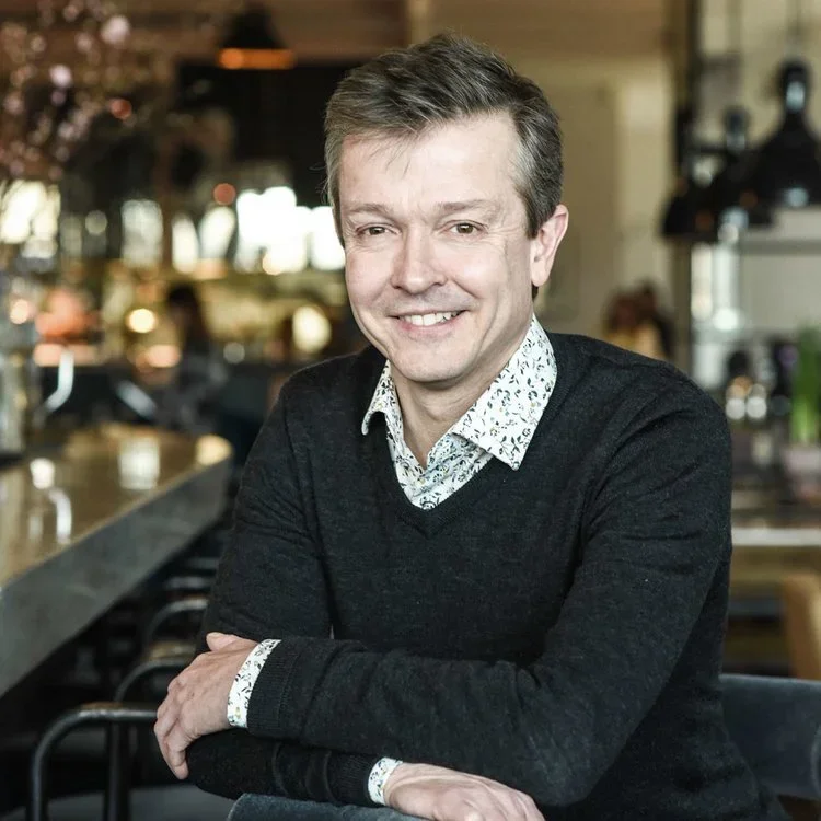 A smiling man with light brown hair wearing a white patterned shirt and dark sweater, sitting with arms crossed at a bar or restaurant.