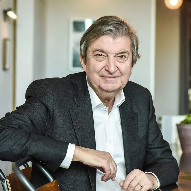 Portrait of an older man with gray hair in a white shirt and black blazer, sitting at a table in an indoor setting.