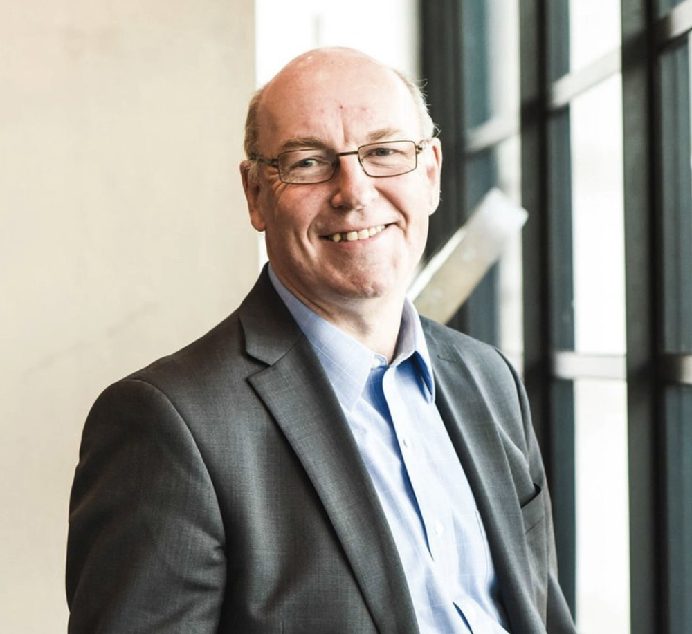 A middle-aged man in a gray suit and glasses smiling, standing near large windows in a modern building.