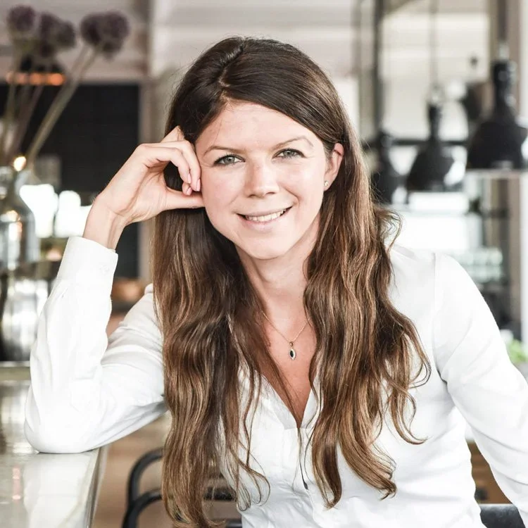 A woman with long brown hair and gray eyes, wearing a white blouse, smiling while sitting in a modern cafe or restaurant with a blurred background of flowers and kitchen equipment.