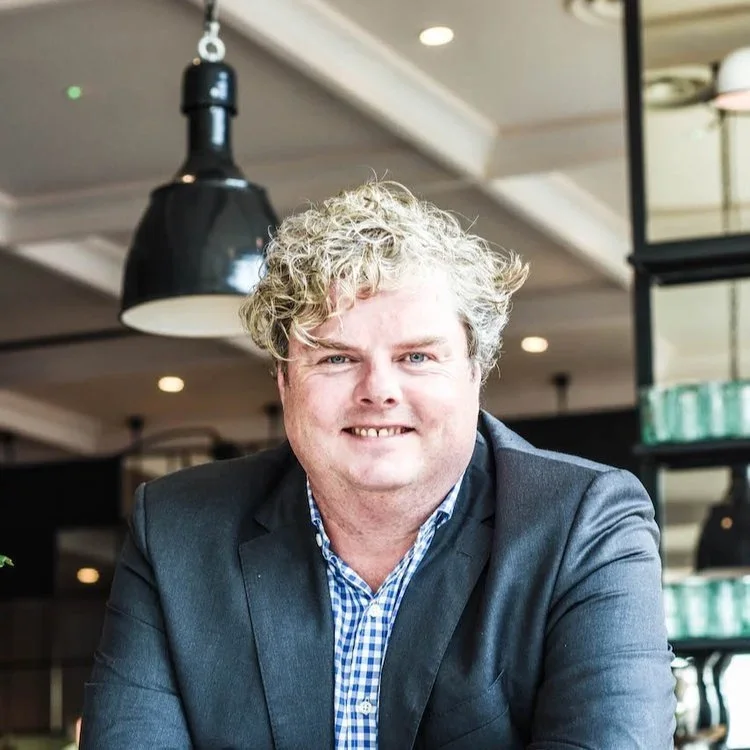 A middle-aged man with curly blond hair wearing a dark blazer and blue checkered shirt, smiling at the camera inside a well-lit indoor setting.