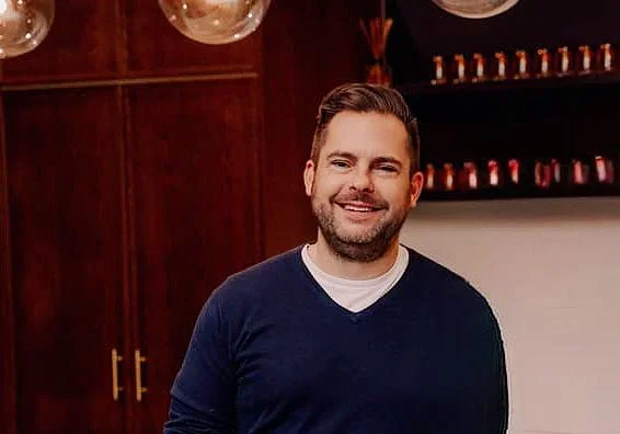 A smiling man with short brown hair and a beard, wearing a navy blue sweater over a white shirt, standing in a room with wooden cabinets and shelves with small jars or containers in the background.
