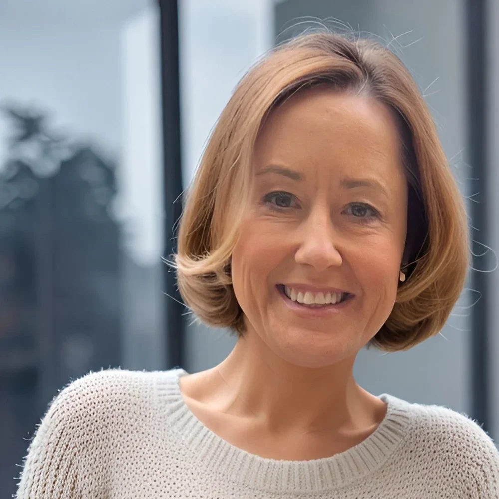 Close-up of smiling woman with short blonde hair, wearing a beige sweater, in front of a blurred outdoor background with buildings and trees.