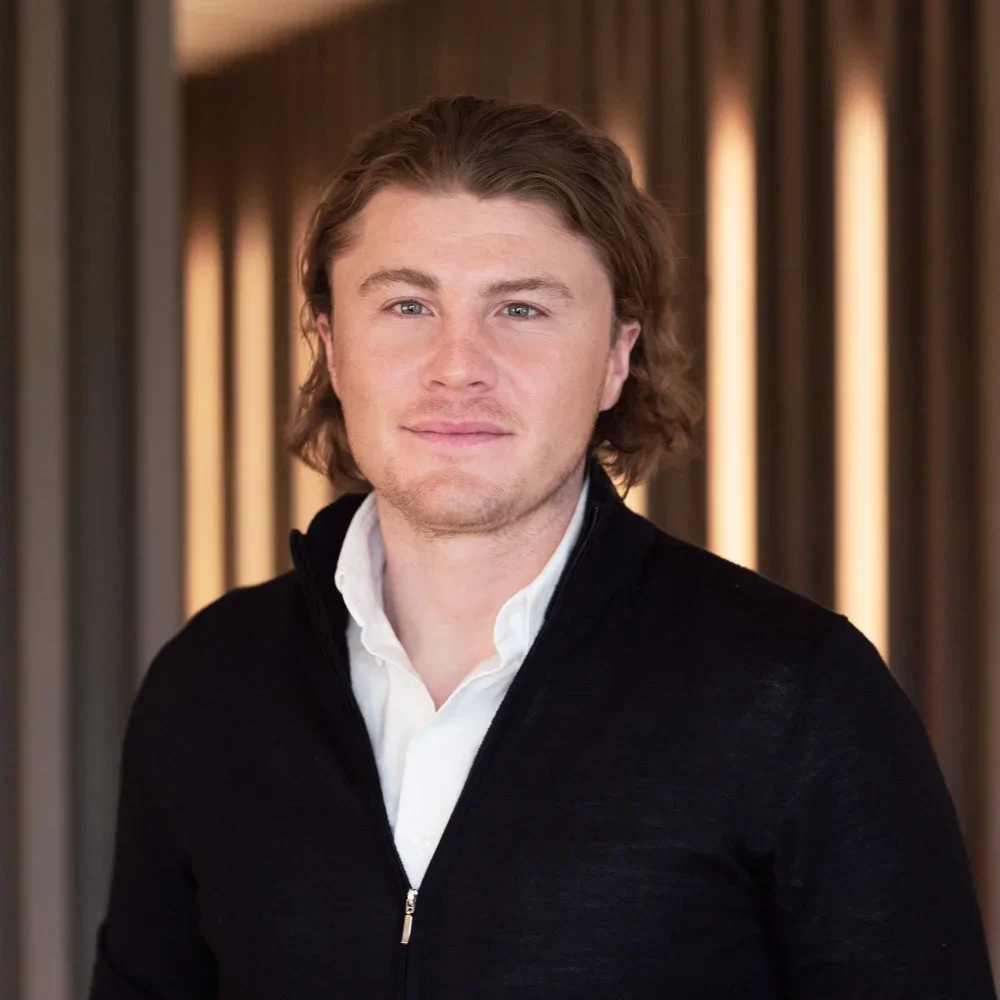 A young man with shoulder-length light brown hair and a light beard, wearing a white collared shirt and a black zip-up sweater, standing in front of a backdrop with vertical wooden panels.