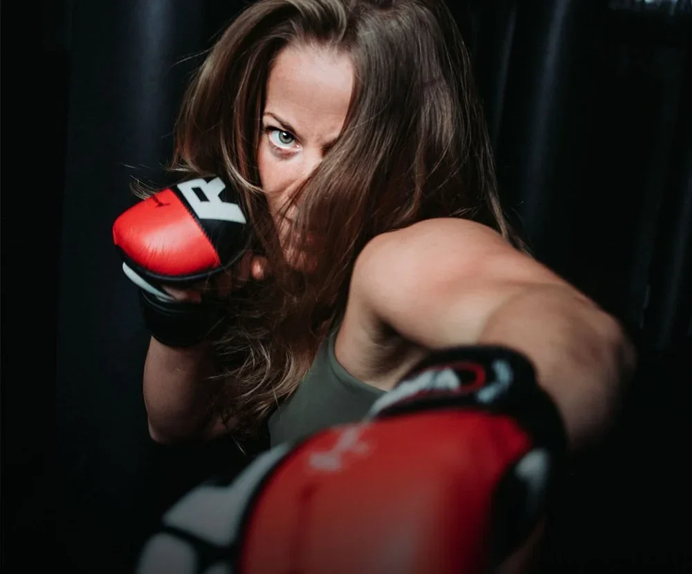 A woman with long brown hair wearing red and black boxing gloves, throwing a punch towards the camera in a fighting stance.