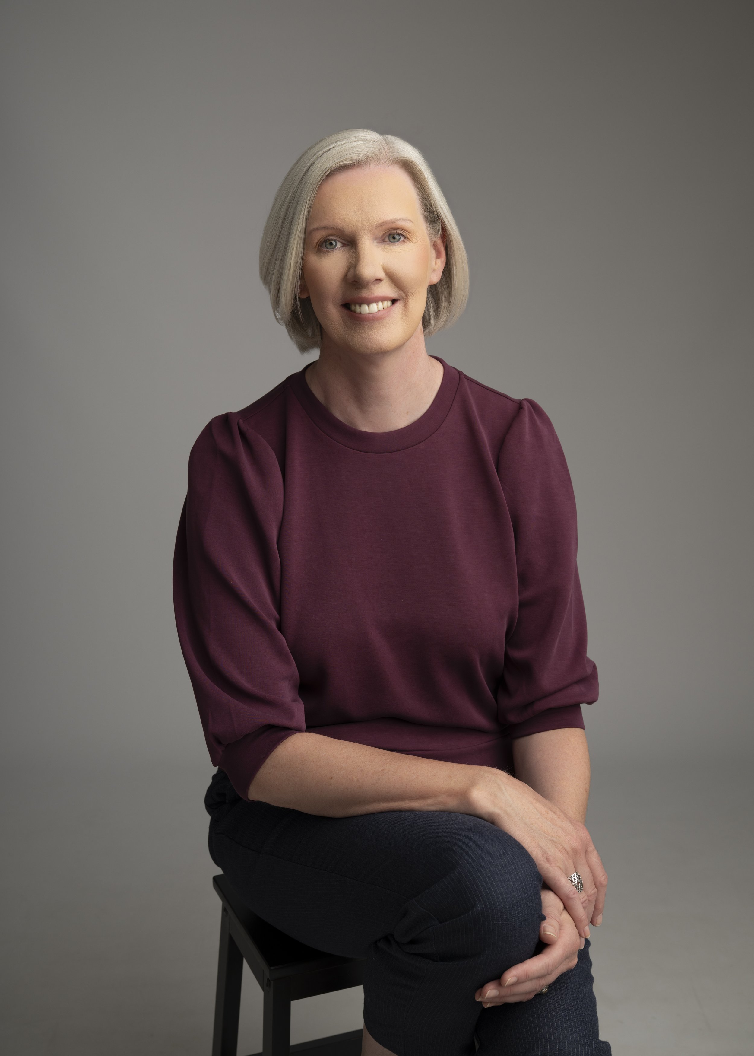 A woman with short gray hair, wearing a maroon top with puffed sleeves, sitting on a black stool against a plain gray background, smiling at the camera.