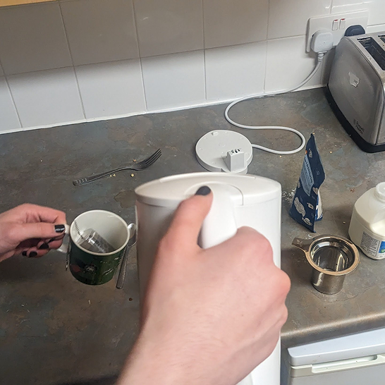 A hand holding a kettle over a mug with a metal loose-leaf infuser.