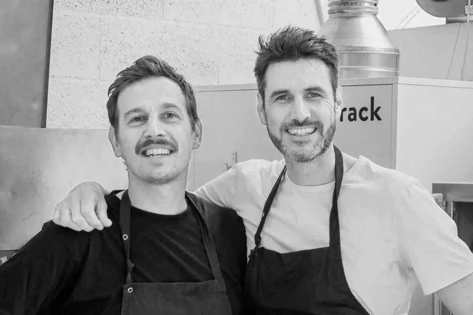 Two smiling men wearing aprons posing together in a kitchen.