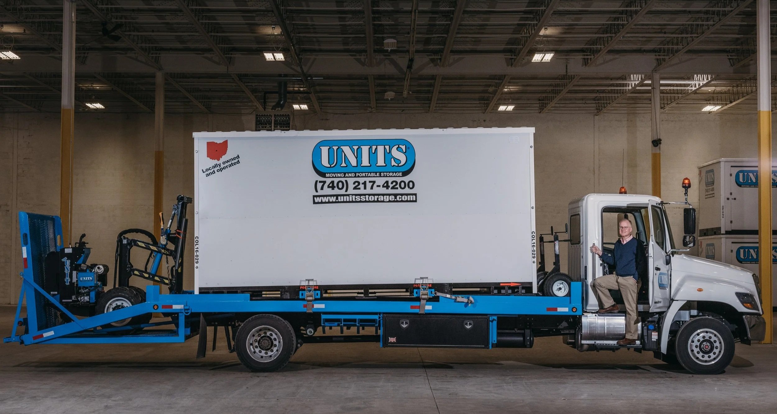 A man sitting in the driver's seat of a moving truck, which is parked inside a large warehouse. The truck has a large box with company logo and contact information for 'UNITS Moving and Portable Storage'.