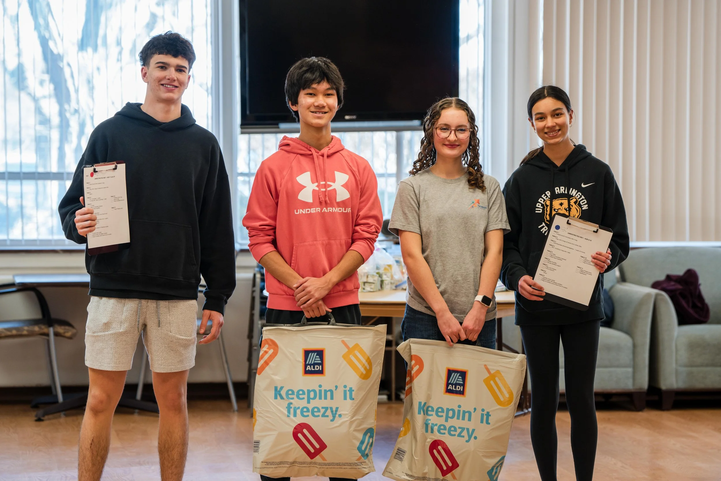 Four teenagers standing indoors holding ALDI shopping bags and clipboards, some smiling.
