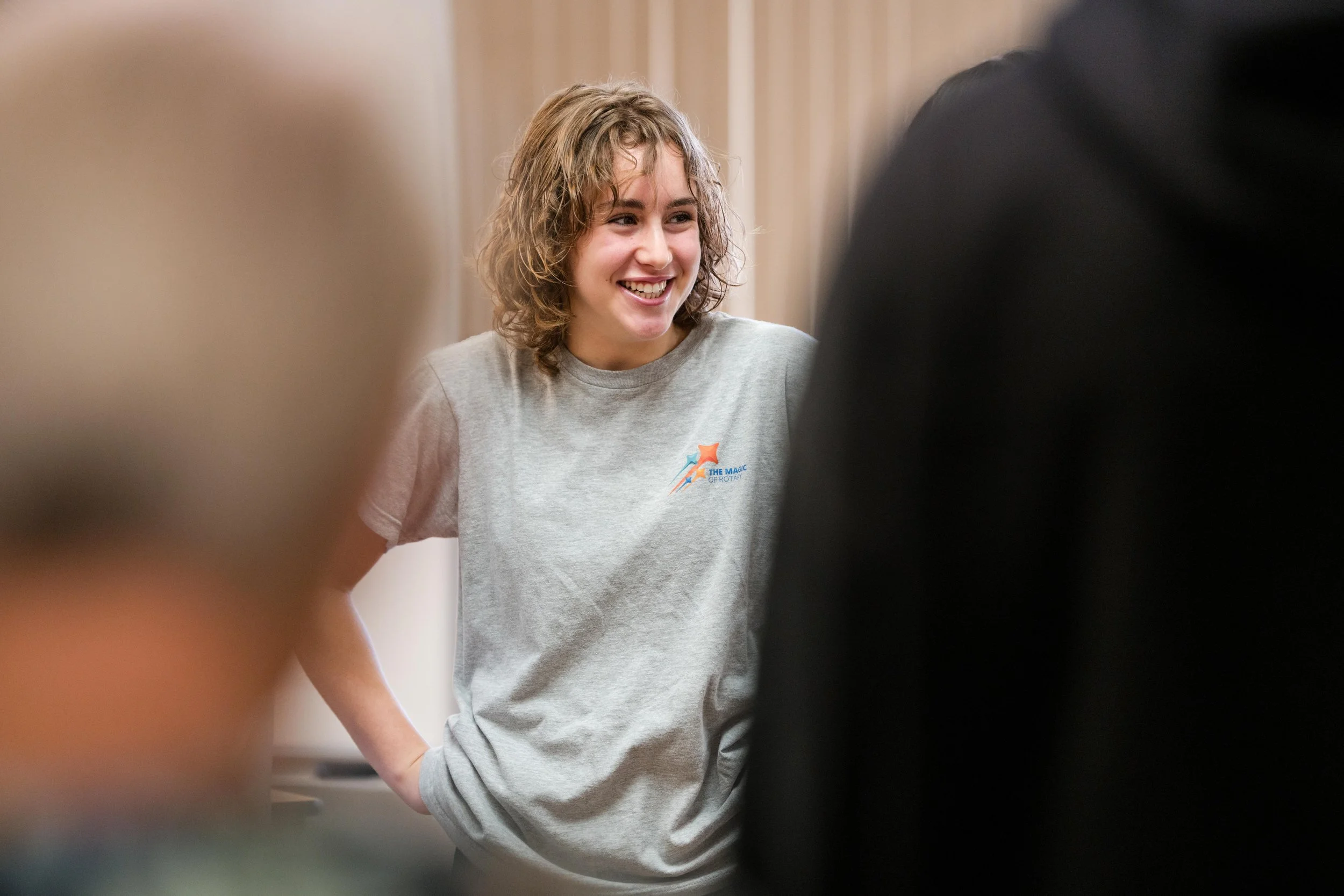 A smiling young woman with curly hair wearing a gray T-shirt with an orange and blue logo, standing indoors and talking to others.
