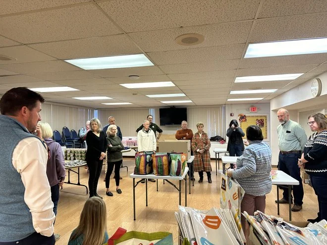 Group of people standing and sitting in a community room with tables and chairs, some looking at a woman speaking at a table with various wrapped gifts or items on display.