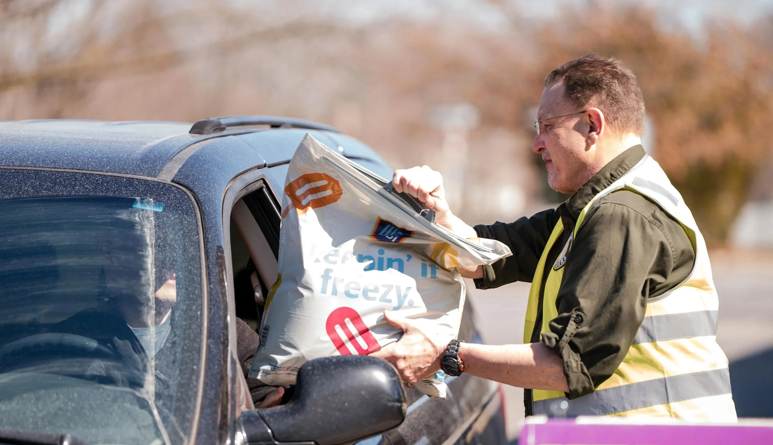 Man handing a large bag of frozen goods to a driver through the open car window on a sunny day.