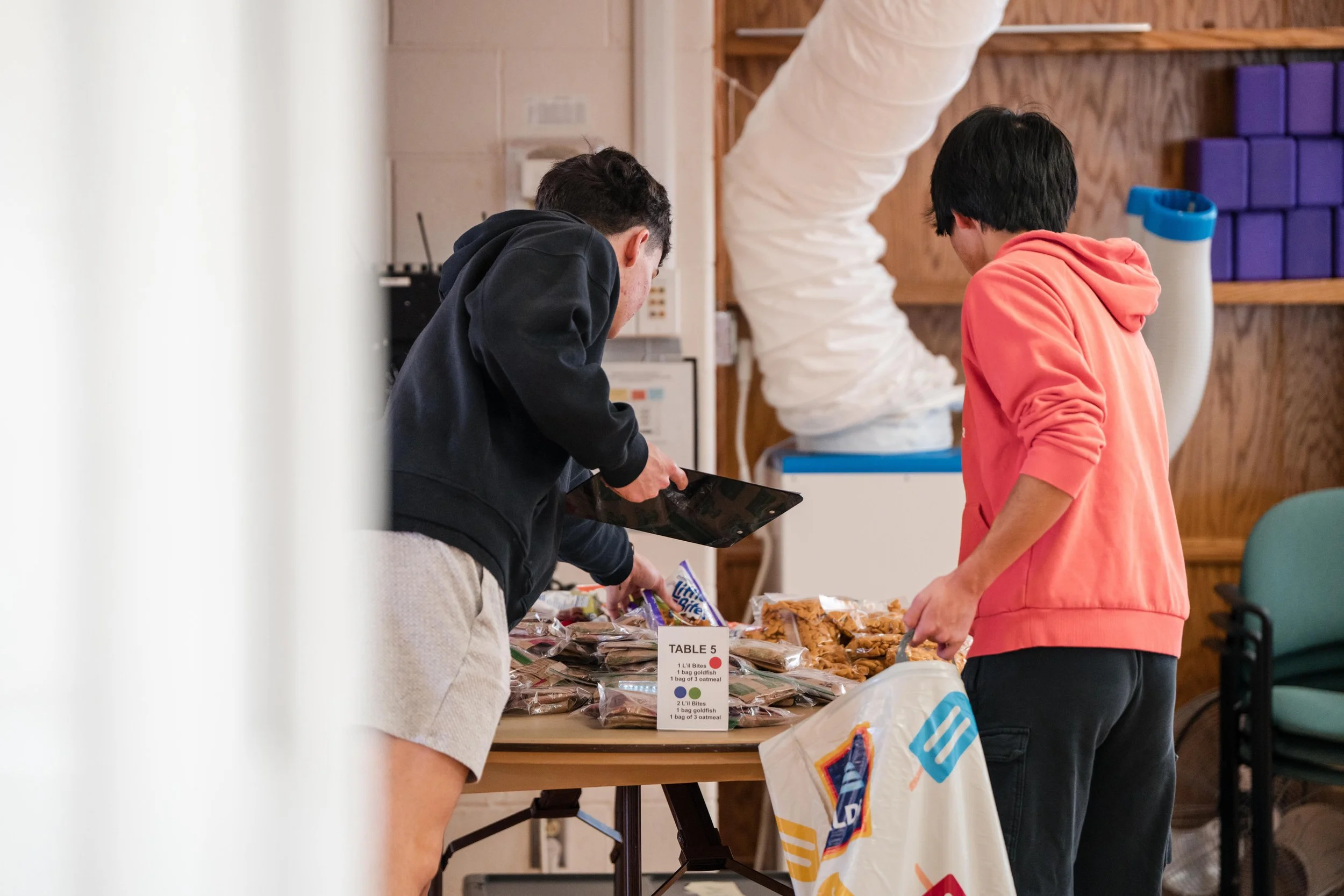 Two young men browsing snacks at a table in a room with wooden walls, with purple blocks and a large white hose in the background.