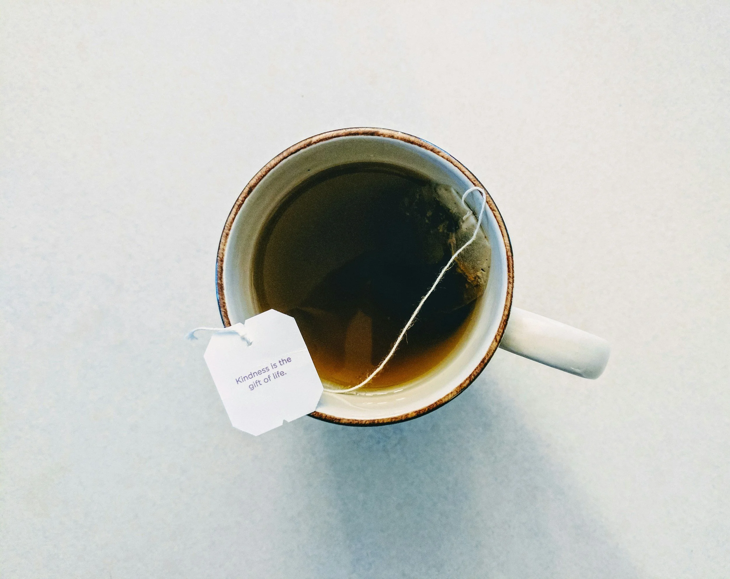 Top-down view of a white teacup with a brown rim, filled with hot tea and a tea bag inside. A white tag on the tea bag reads "Kindness is the gift of life." Place is on a light-colored surface.