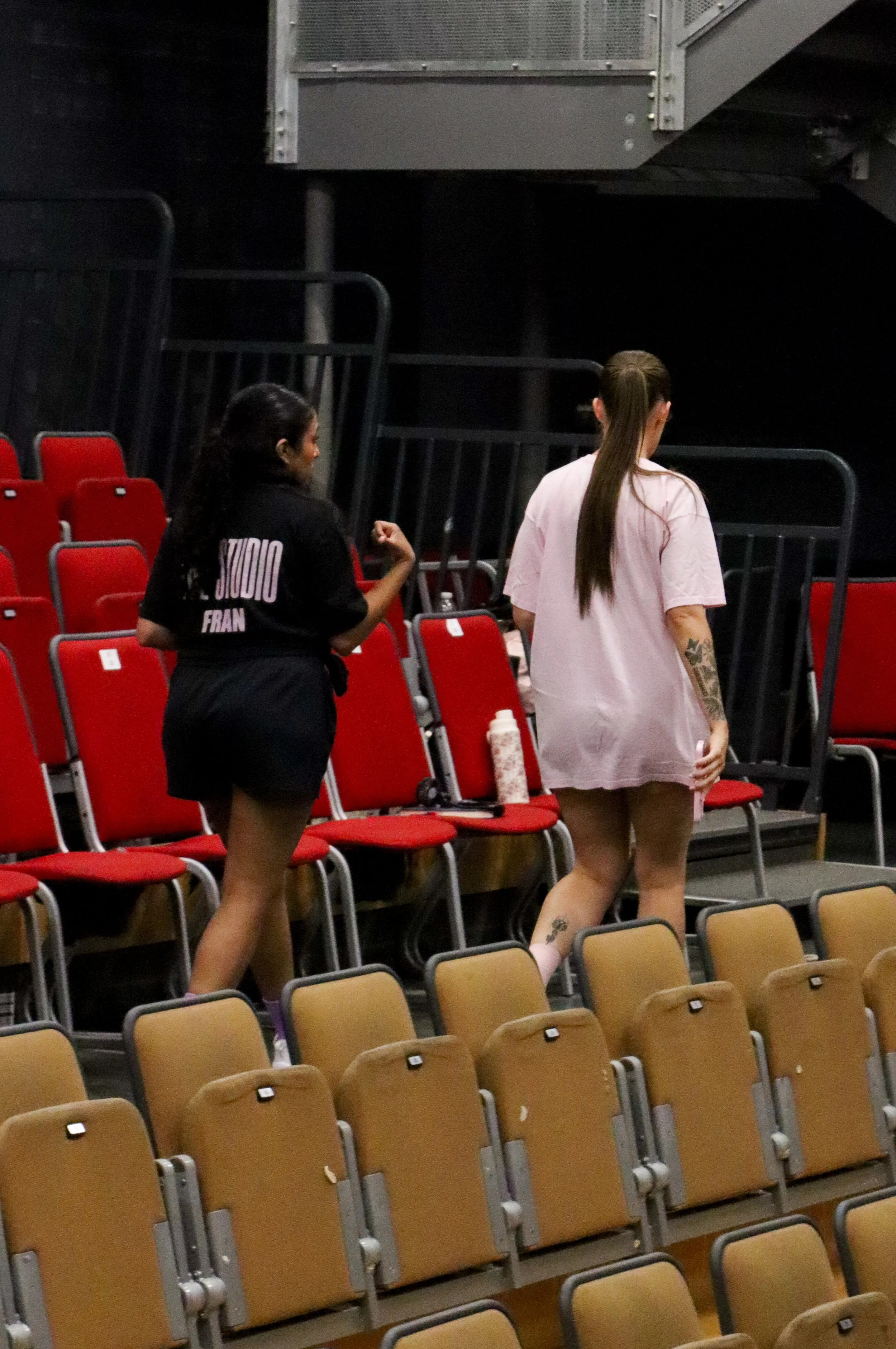 Two women walking through an empty auditorium with red and beige chairs, one wearing a black shirt and shorts, the other wearing a oversized pink T-shirt and shorts.