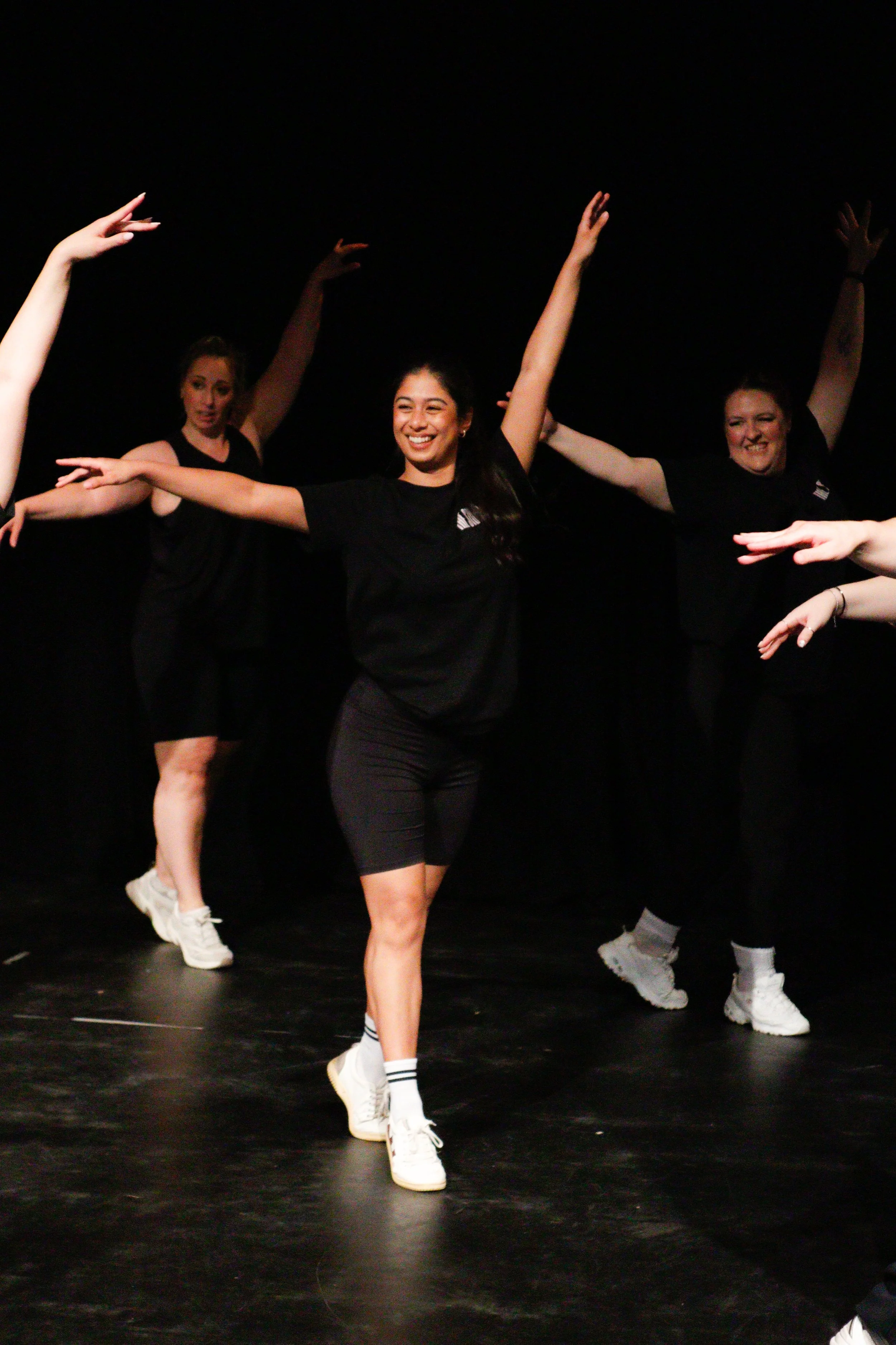 Group of young women dancing on stage, wearing black t-shirts, shorts, and white sneakers, with arms raised and smiling.
