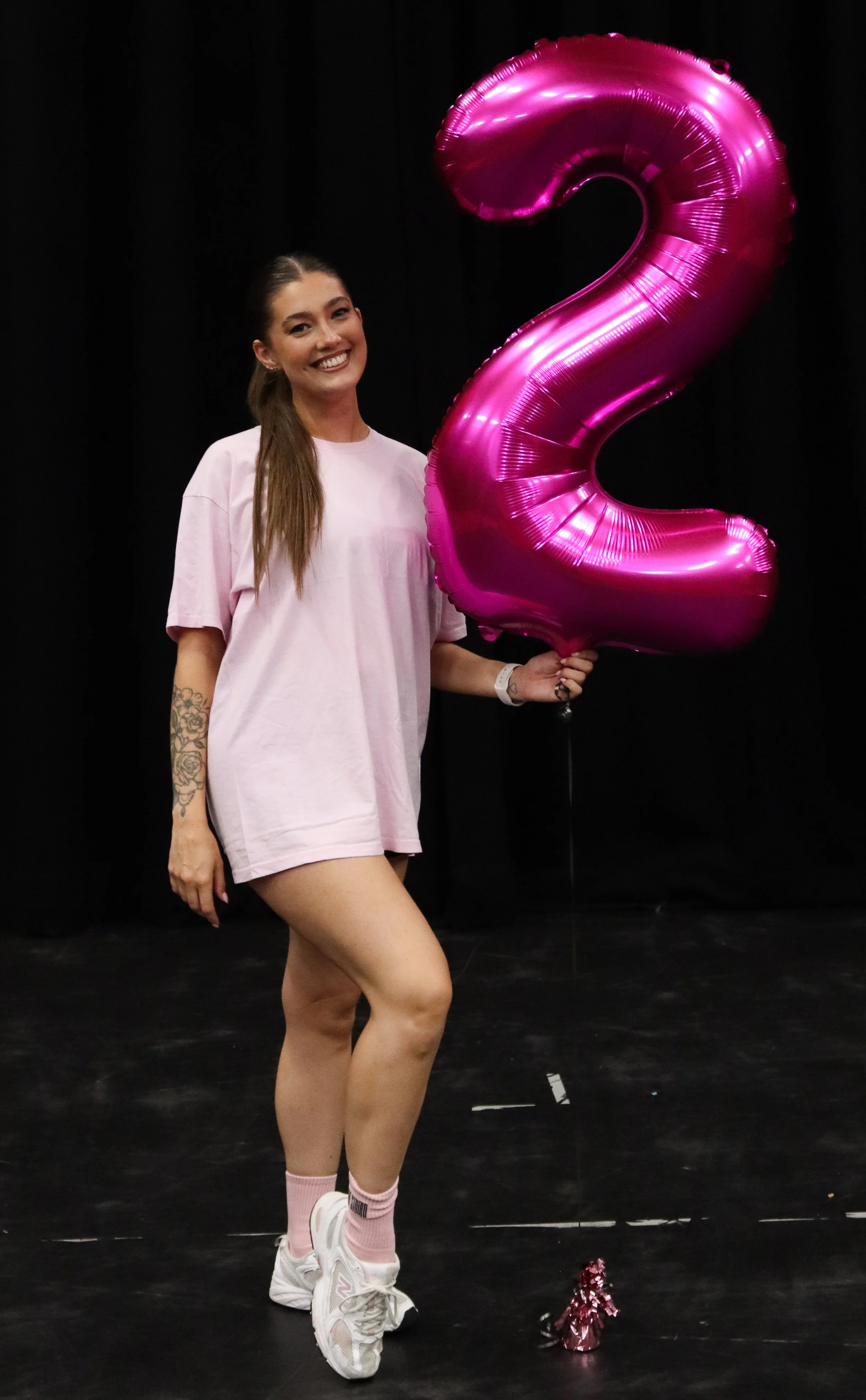 A woman dressed in pink athletic wear holding a large pink number 2 balloon, standing on a black stage with a black background.