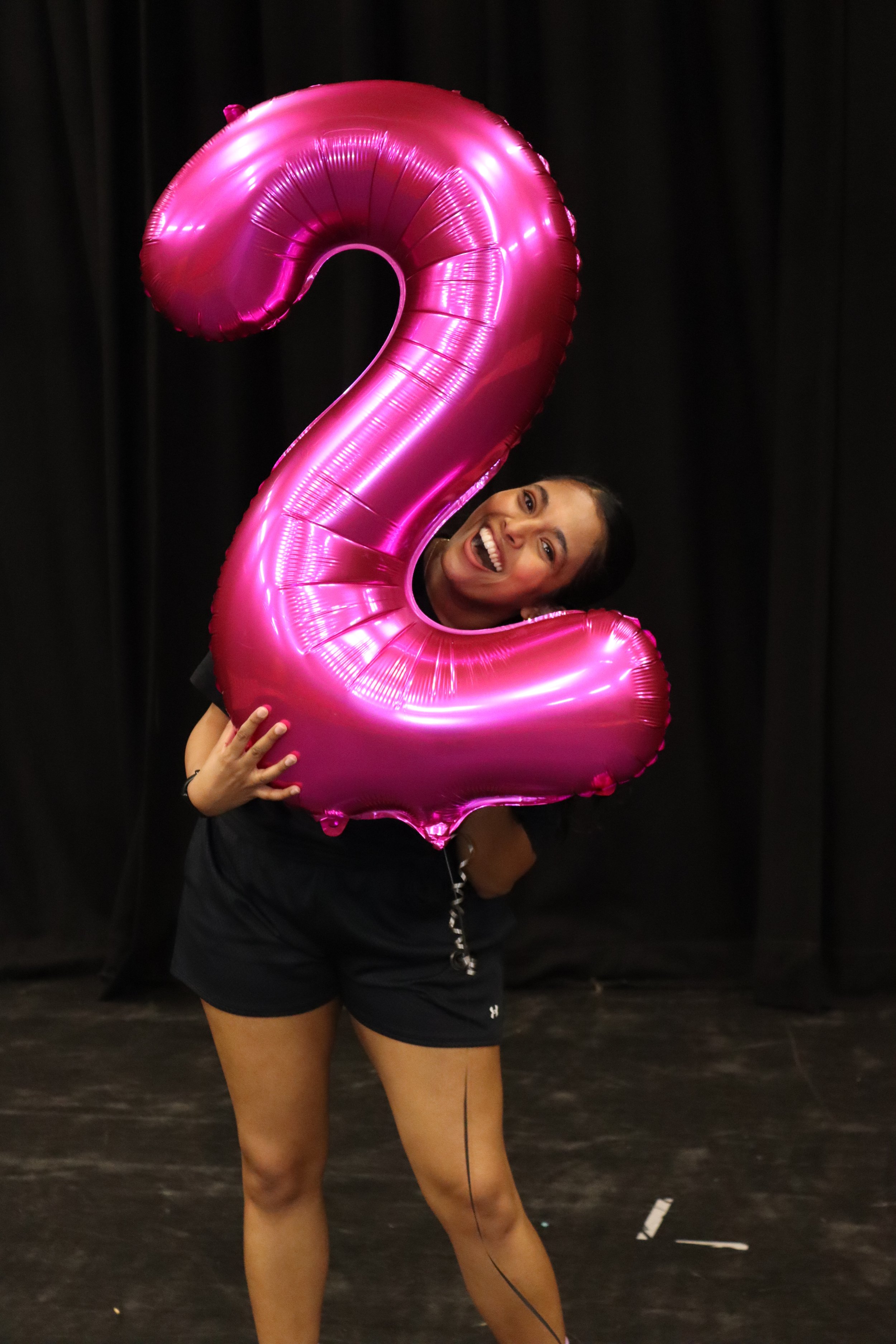 A woman holding a large, pink, number two-shaped balloon in front of a black background.