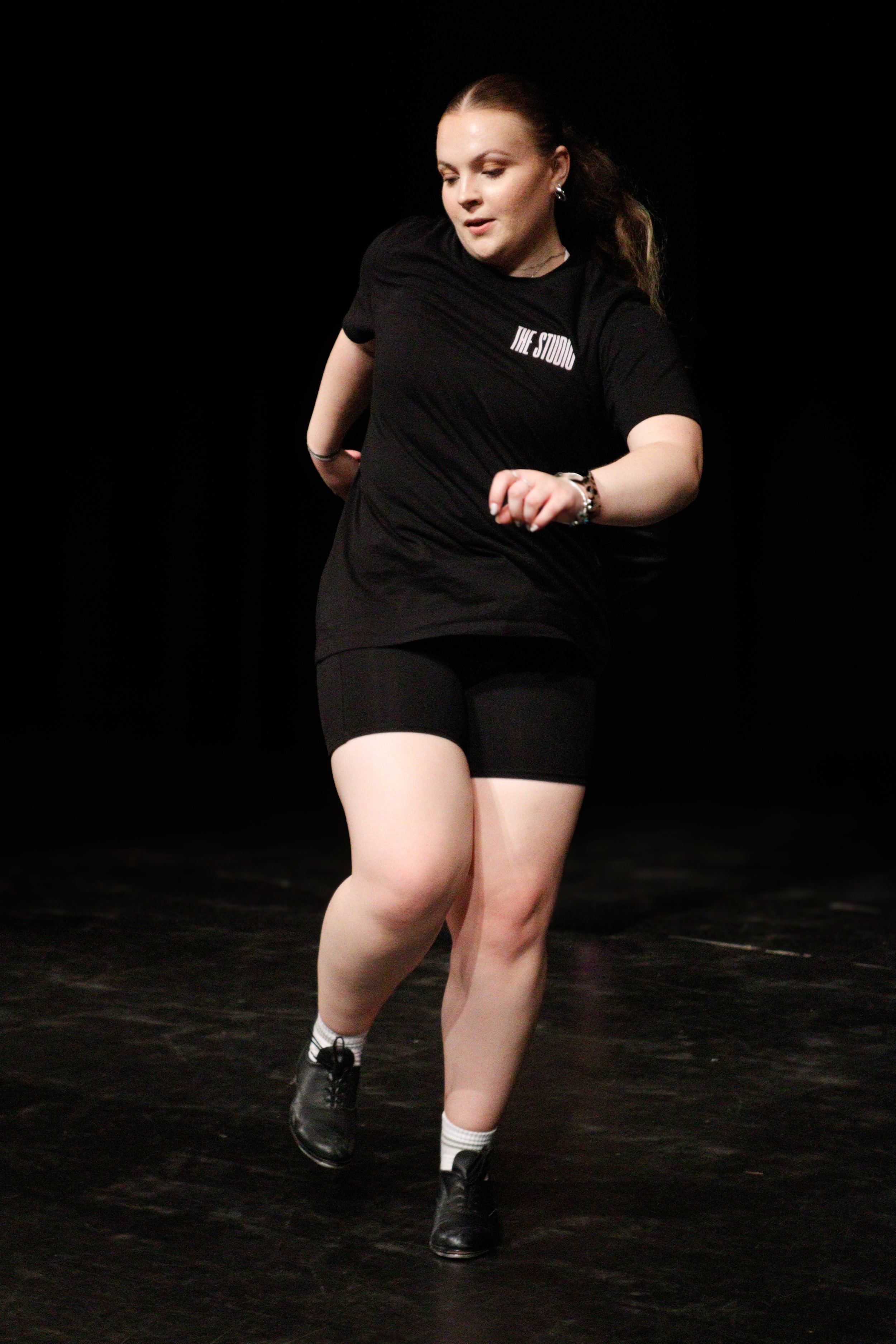 A woman with red hair dances on a dark stage, wearing a black t-shirt that says 'THE STUDIO,' black shorts, and black shoes with white socks.