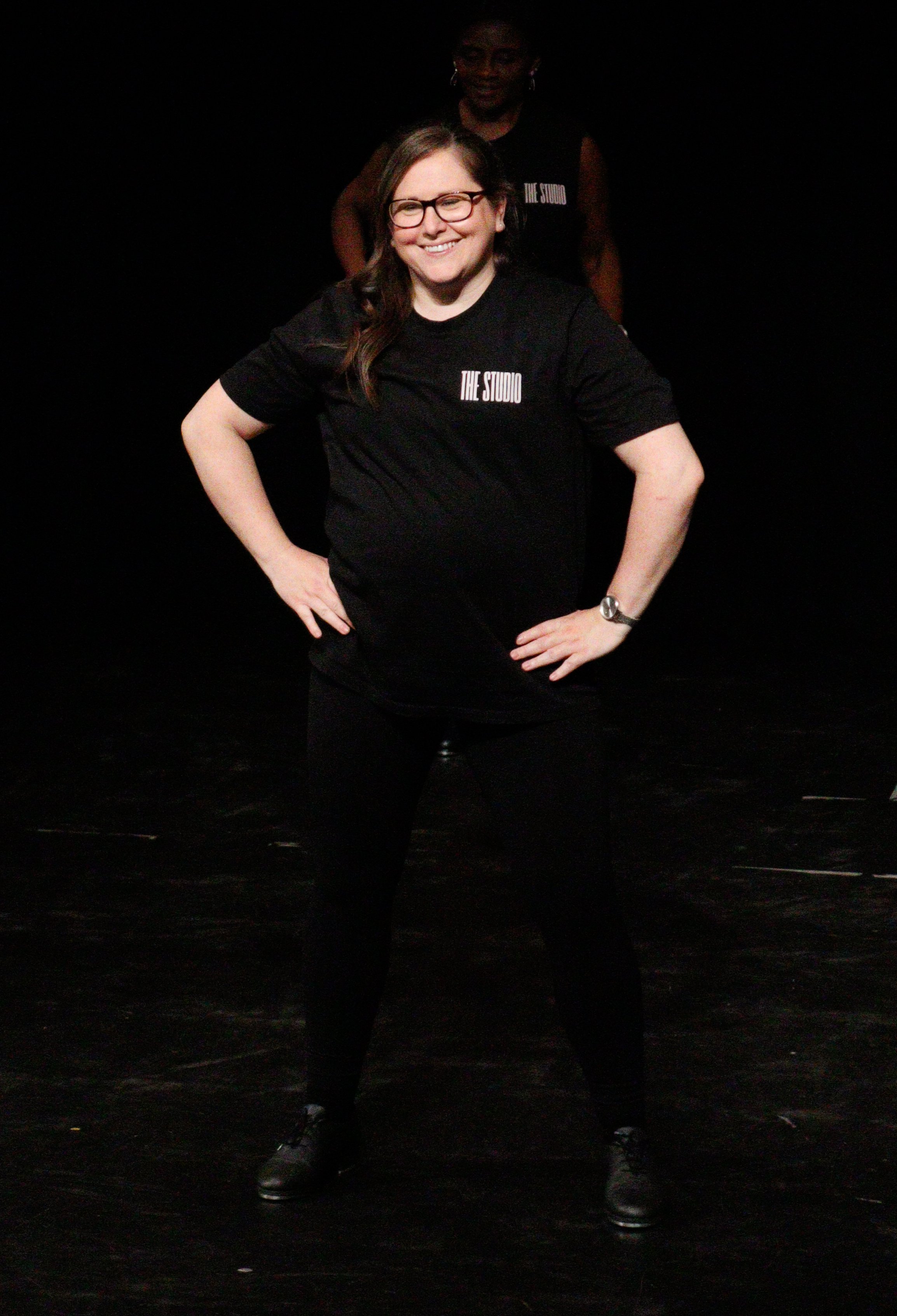 A woman with glasses smiling, standing with hands on hips on a dark stage, wearing a black t-shirt that says 'The Studio', with another person in the background also wearing a similar shirt.