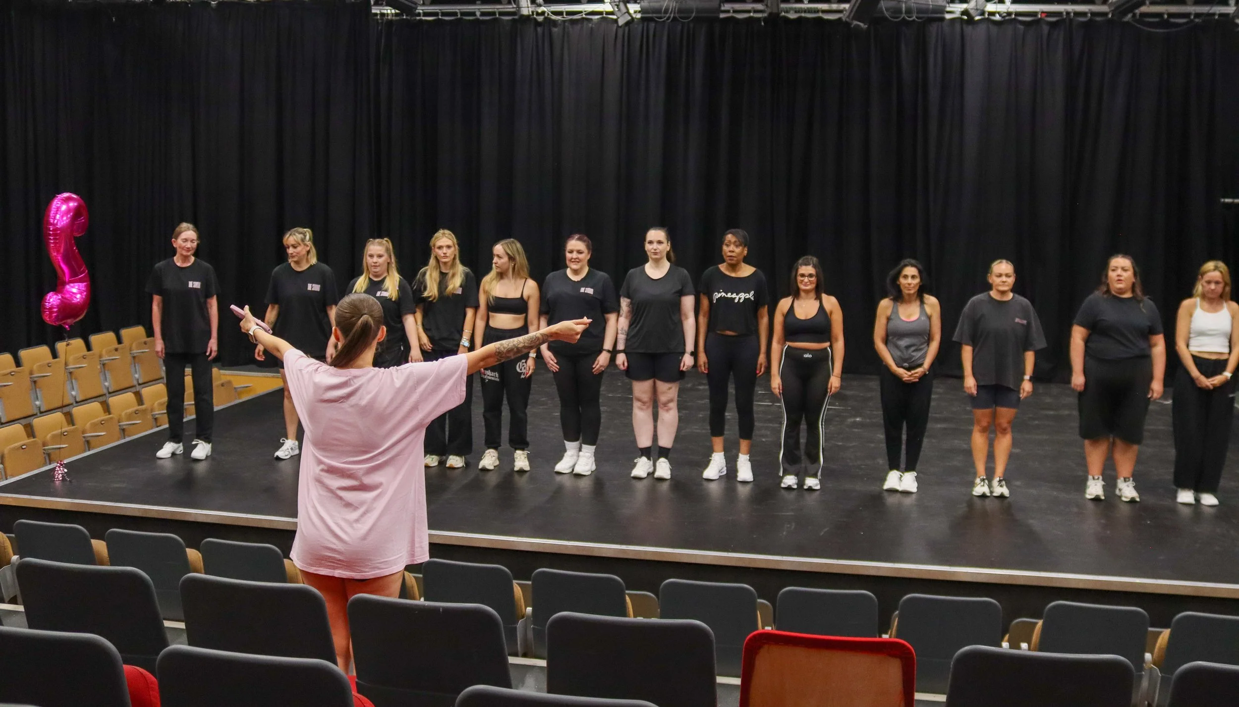 A group of women standing on a stage in front of a black curtain, being directed by a woman in the foreground with tattooed arms, wearing a pink shirt. The stage has black flooring, and there are empty beige chairs and a large pink balloon in the sha