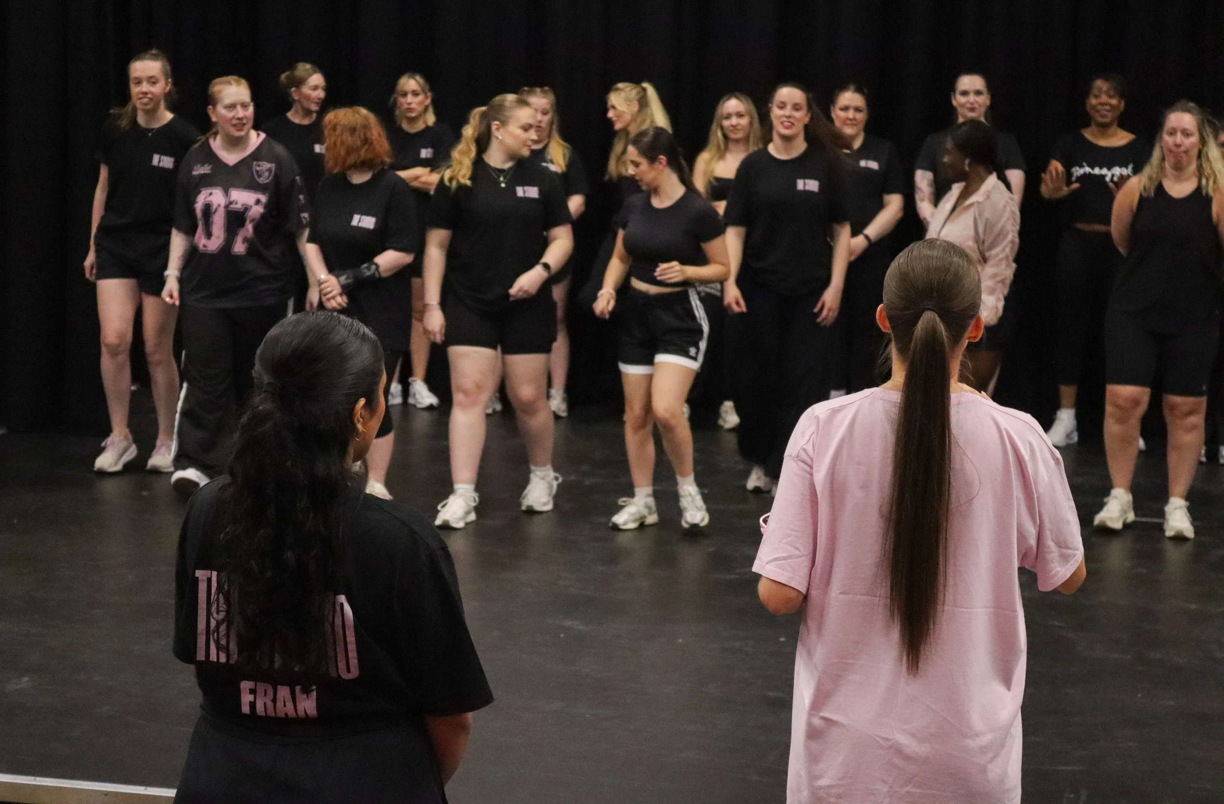 Group of women standing on stage, some are smiling, wearing black clothing, with one woman in a pink jacket and another woman in a black sports uniform, with an audience member with long dark hair in the foreground.