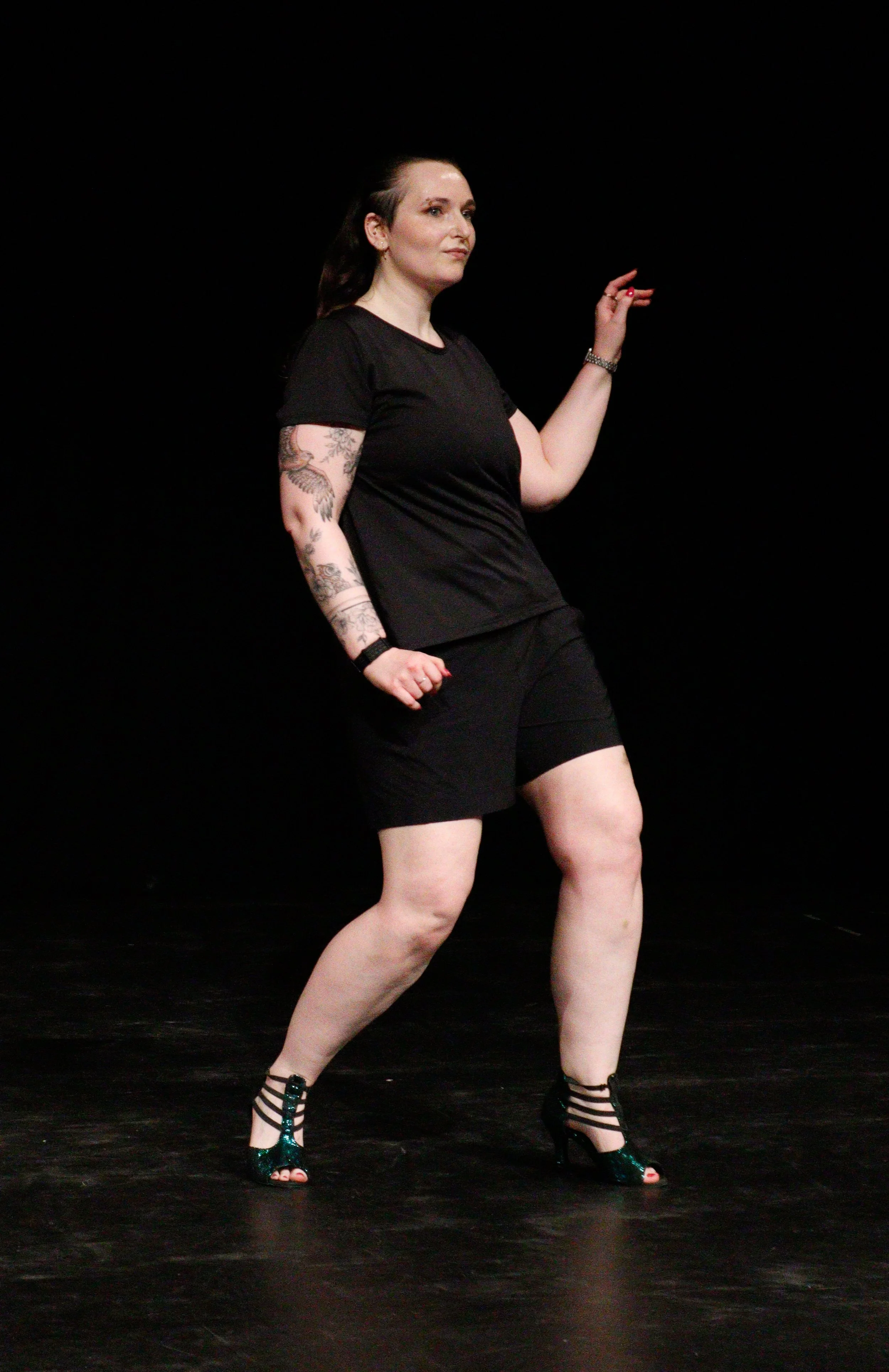 A woman with tattoos on her arms wearing a black t-shirt, black shorts, and high heels dancing on a black stage with a dark background.