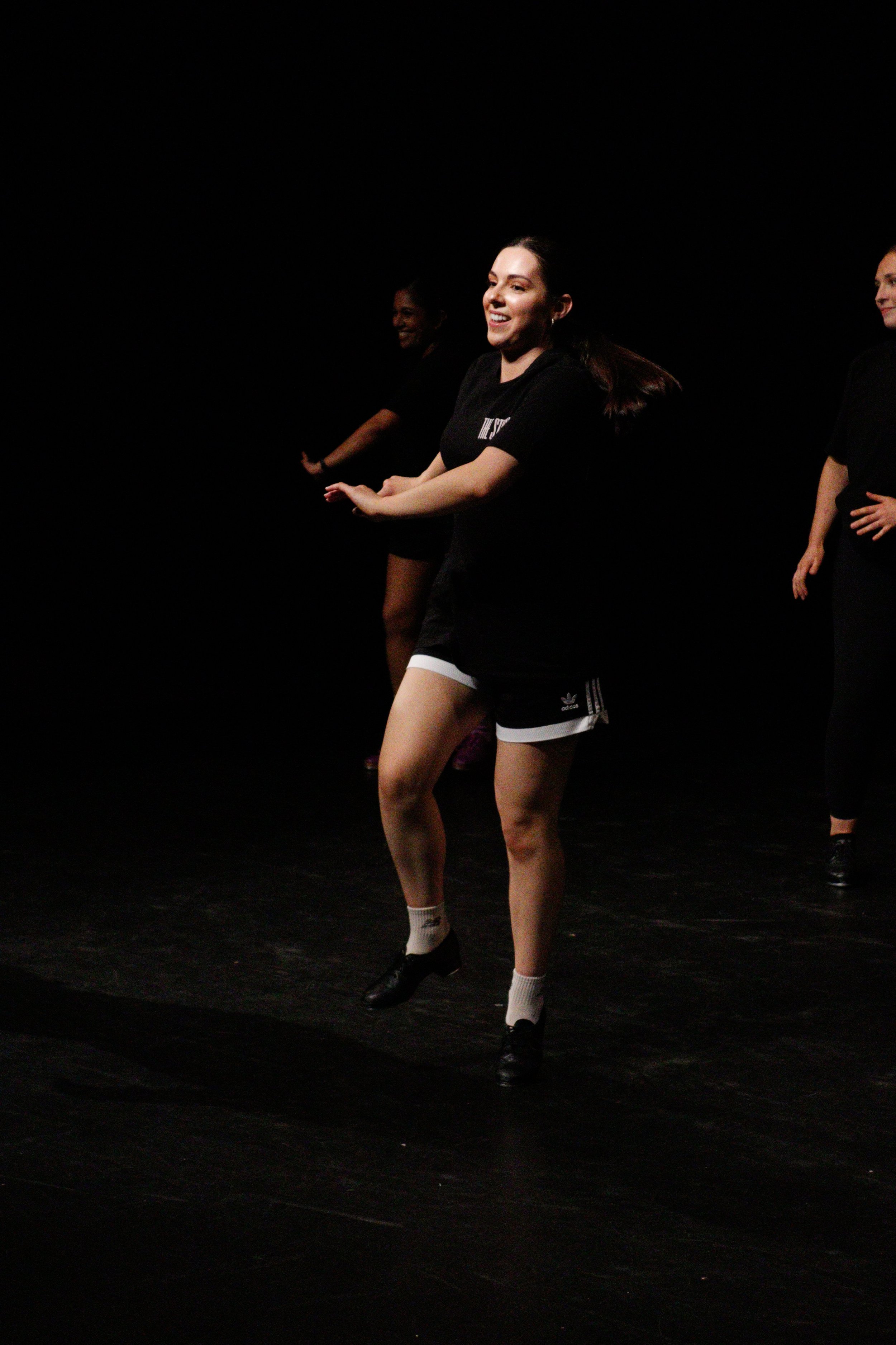 A group of women performing tap dance on a dark stage, wearing black shirts, shorts, and tap shoes.