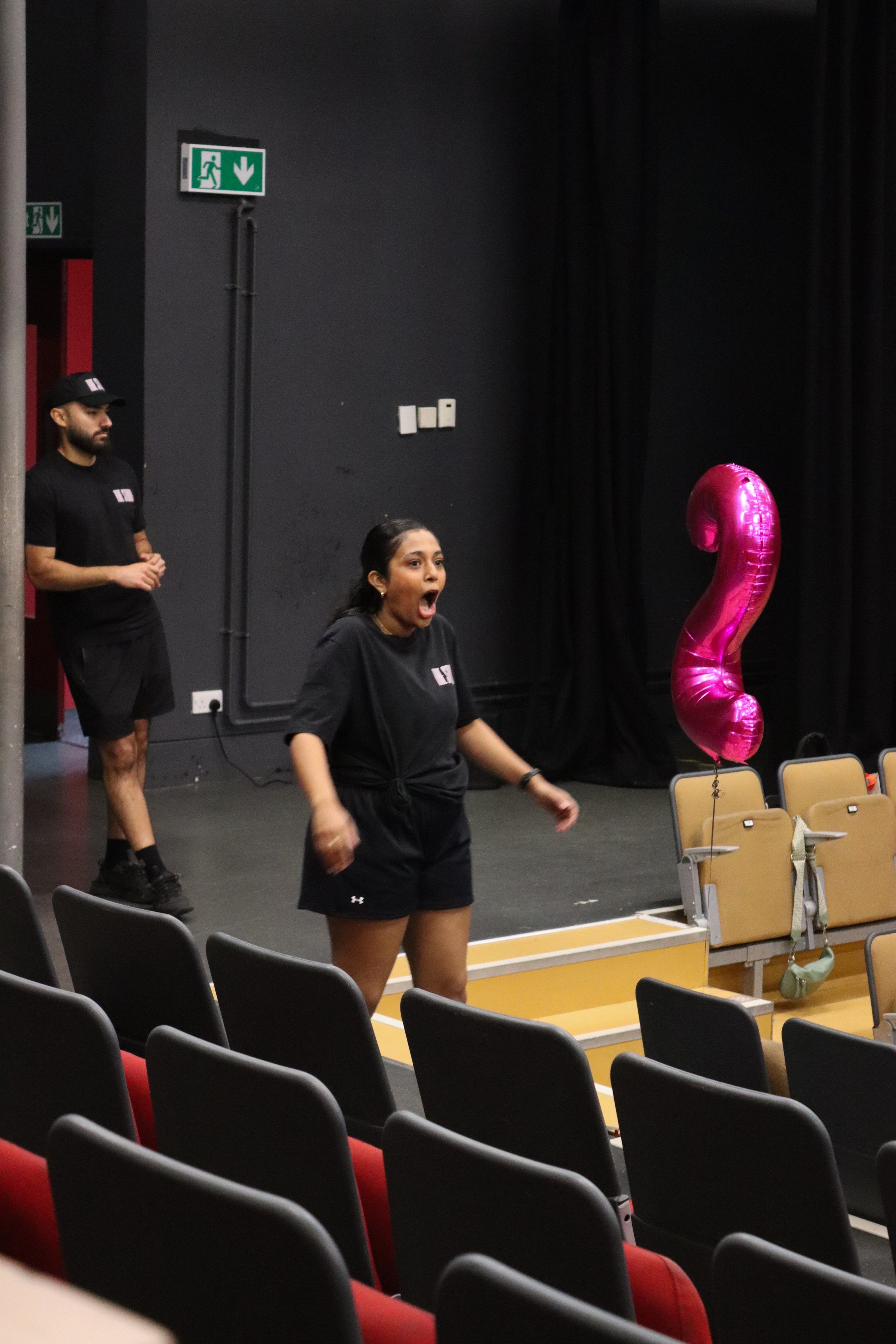 A woman in athletic clothing appears to be surprised or excited, standing in a room with black walls and chairs, near pink foil balloons shaped like the number two, with two people in black shirts in the background.