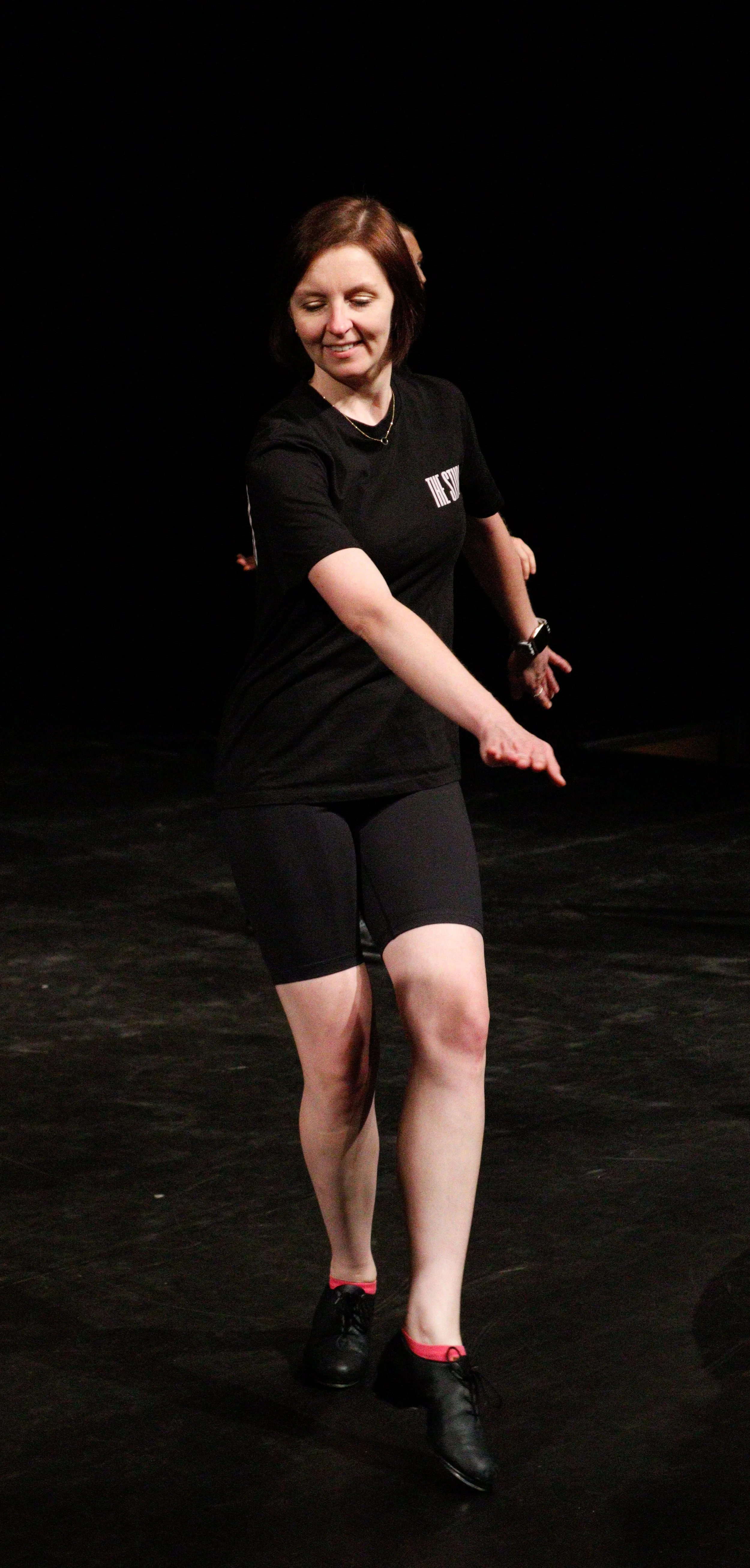 A woman dancing on stage, wearing a black t-shirt, black shorts, and black dance shoes with red accents under her socks.