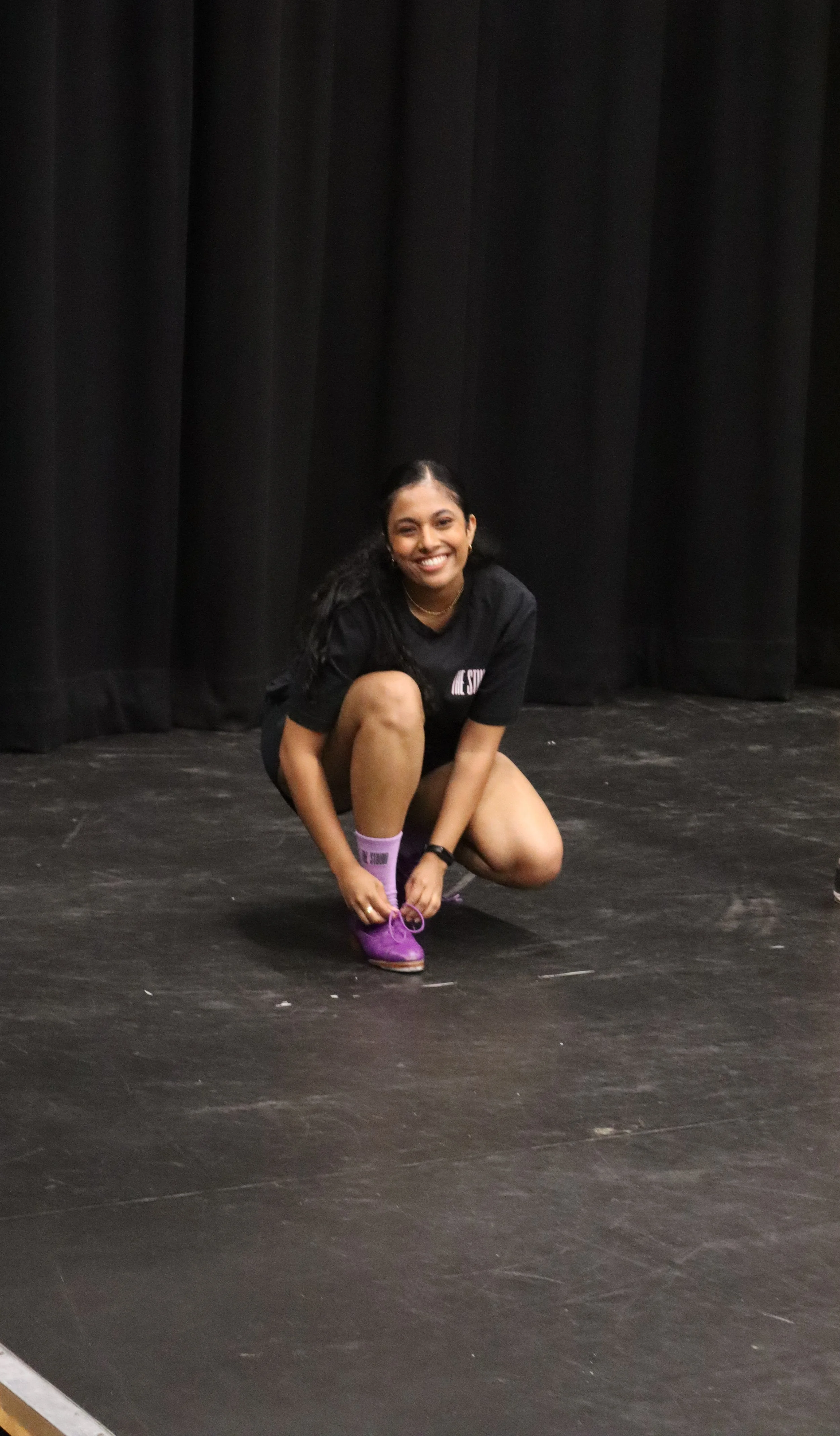 A young woman with long dark hair smiling, crouching and tying her purple shoe on a dark stage with black curtains in the background.