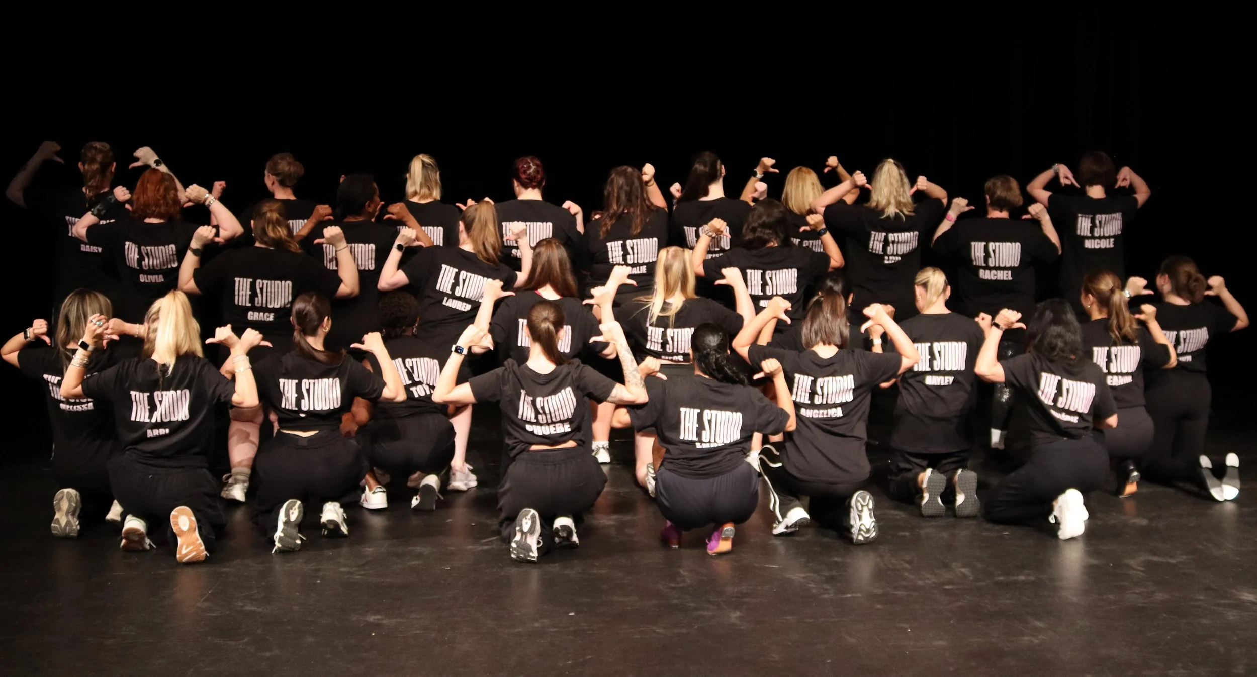 Group of people on stage wearing black T-shirts with 'The Studio' printed on the back, posing with flexed arm muscles.