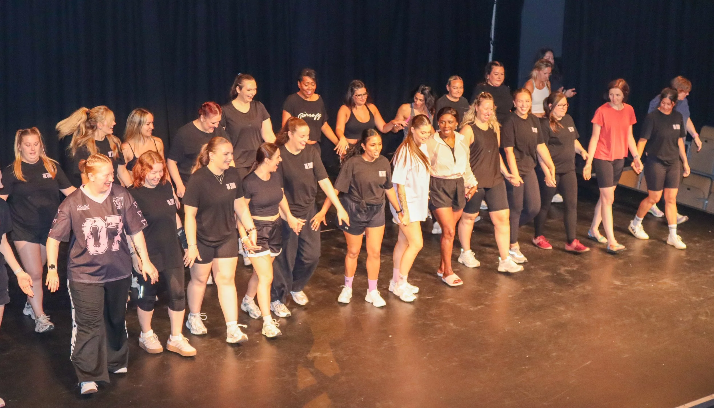 Group of women holding hands and smiling on a stage, participating in a dance or group activity.