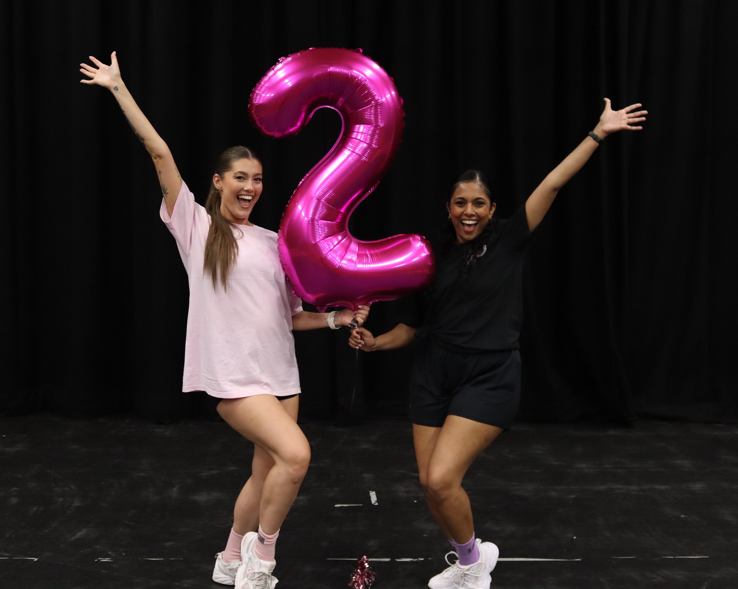 Two women celebrating with a pink metallic balloon shaped as the number 2, on a black stage or background, smiling and dressed casually.