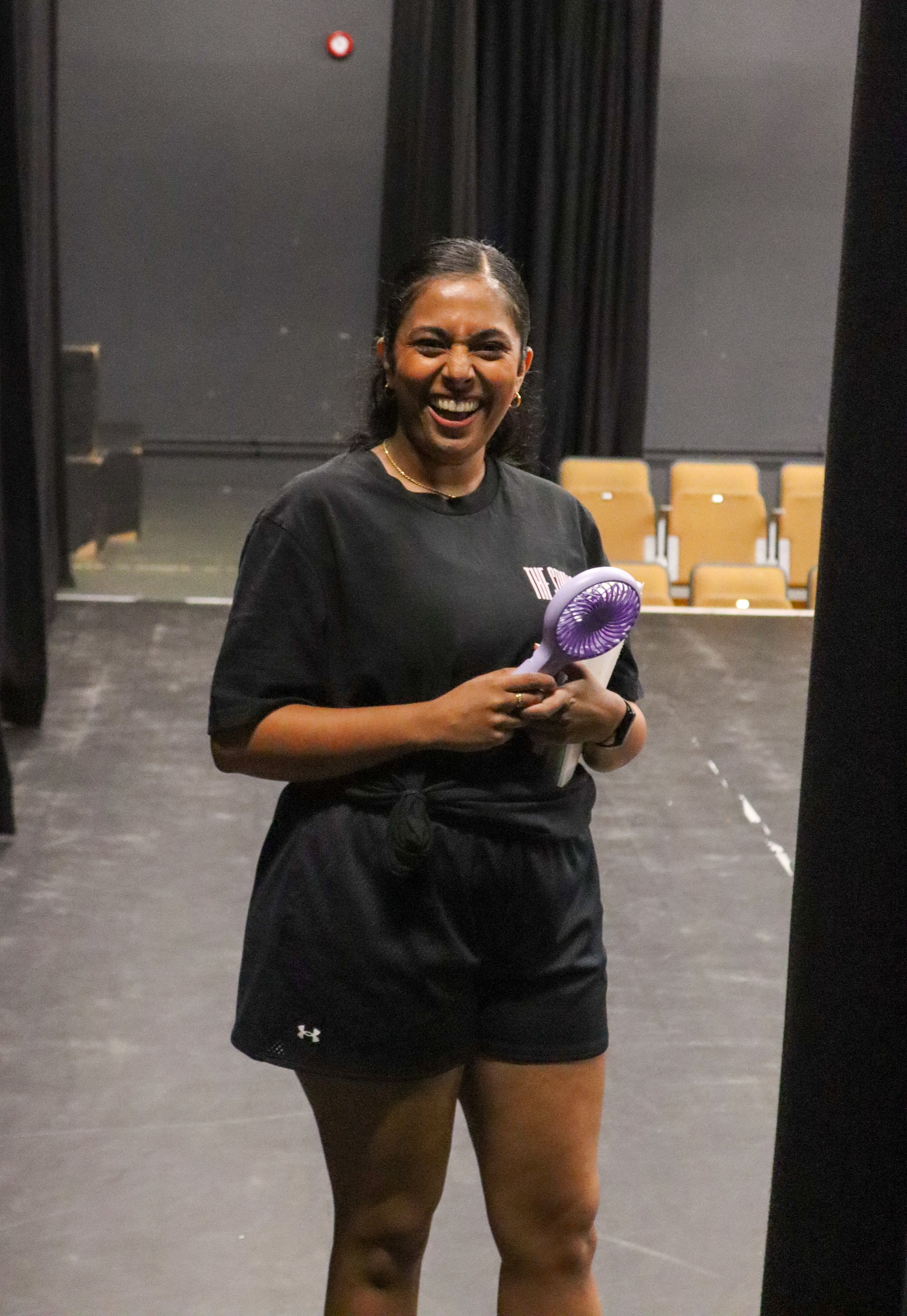 A woman standing on a stage, smiling, holding a purple handheld fan. She is wearing a black t-shirt and black shorts, with black curtains and rows of yellow chairs in the background.