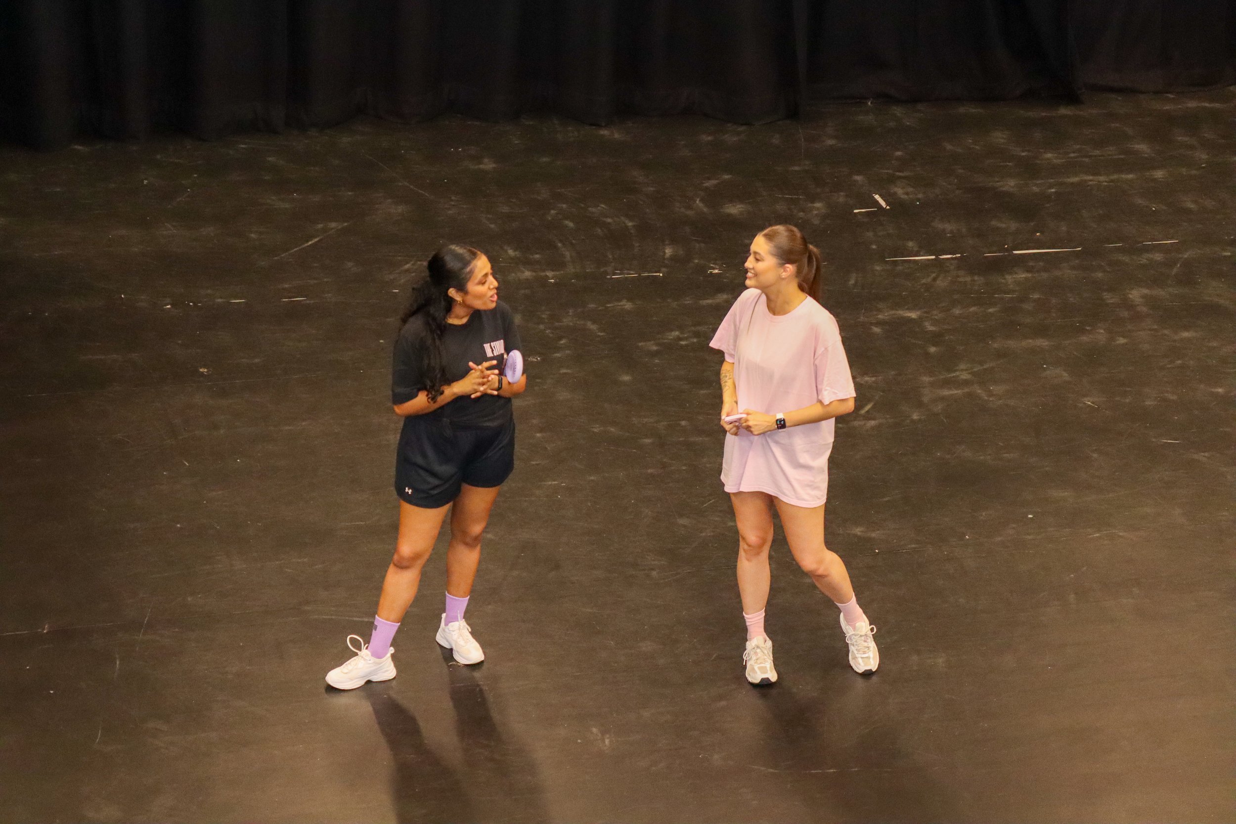 Two women standing apart on a stage, talking to each other. One woman is wearing a black shirt and shorts, and the other is wearing a pink shirt and shorts. The stage has a dark floor and a black curtain in the background.