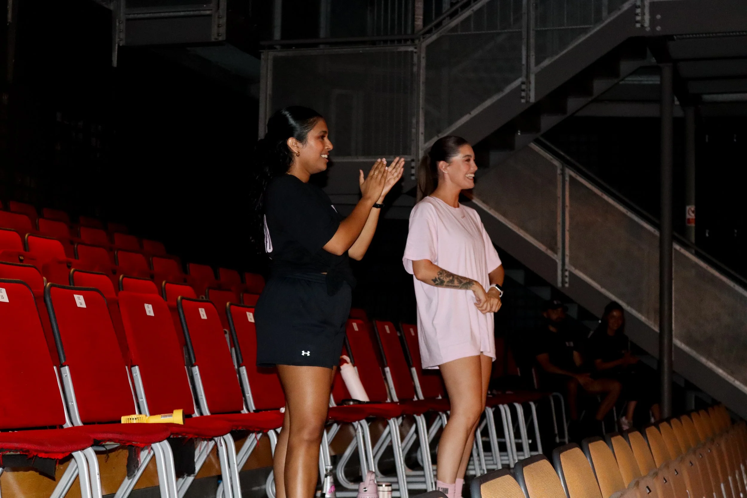 Two women standing in an arena with red seats, clapping and smiling, with industrial stairs and railings in the background.
