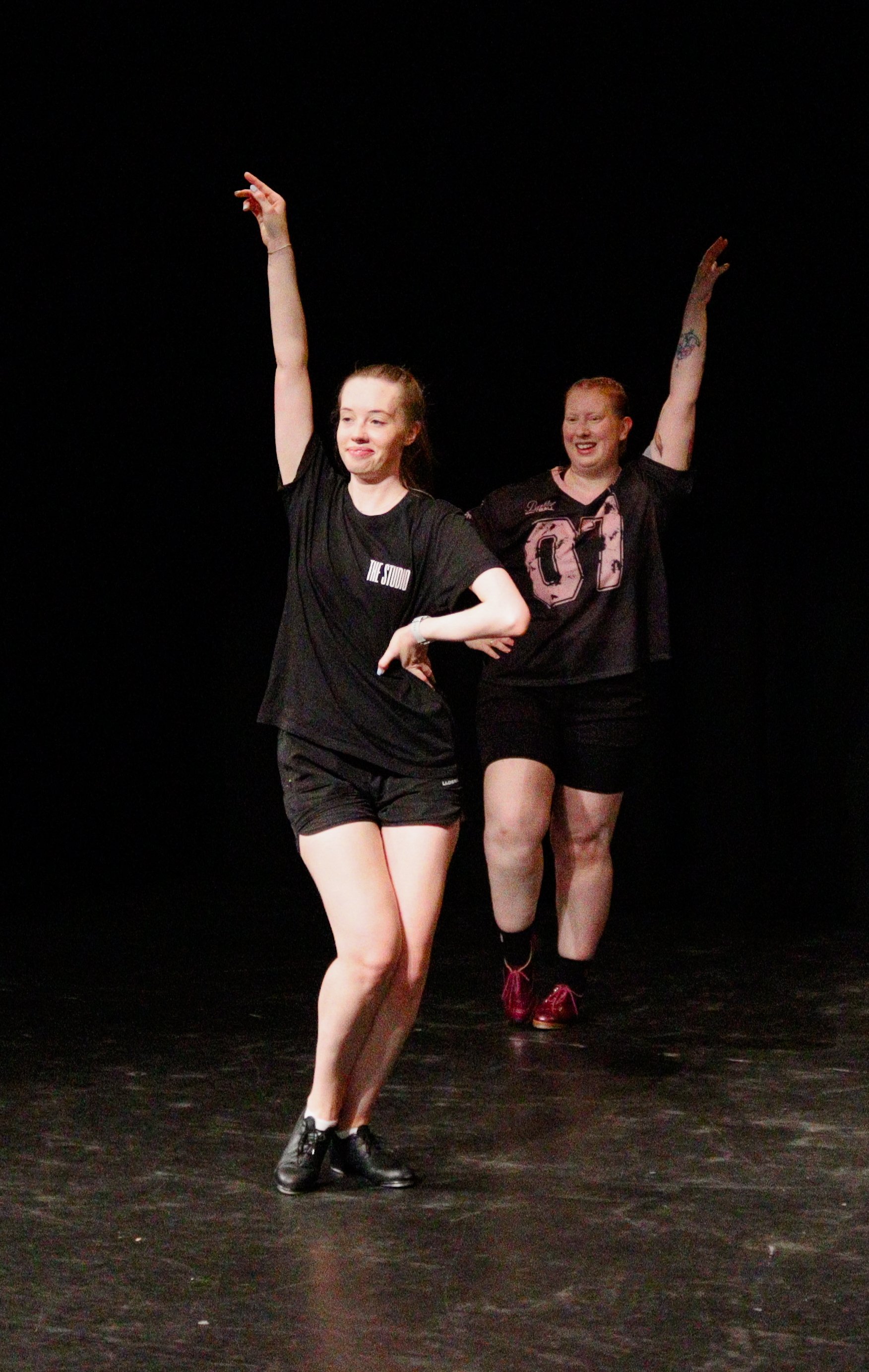 Two women dancing on stage, one in front with a black t-shirt and shorts, and the other behind wearing a sports jersey and shorts, indicating a performance or rehearsal.