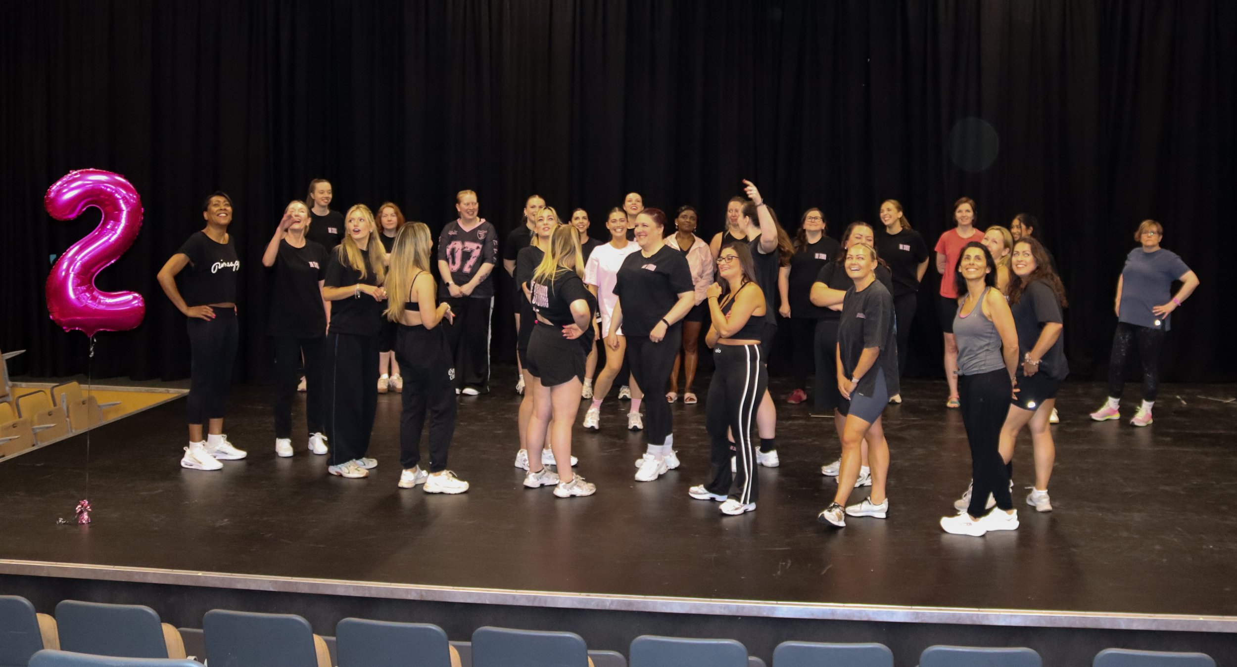 A group of women gather on a stage, celebrating their second anniversary with a large pink balloon shaped like the number two. Some women are smiling and chatting, wearing black workout clothes, while others are looking around or posing.