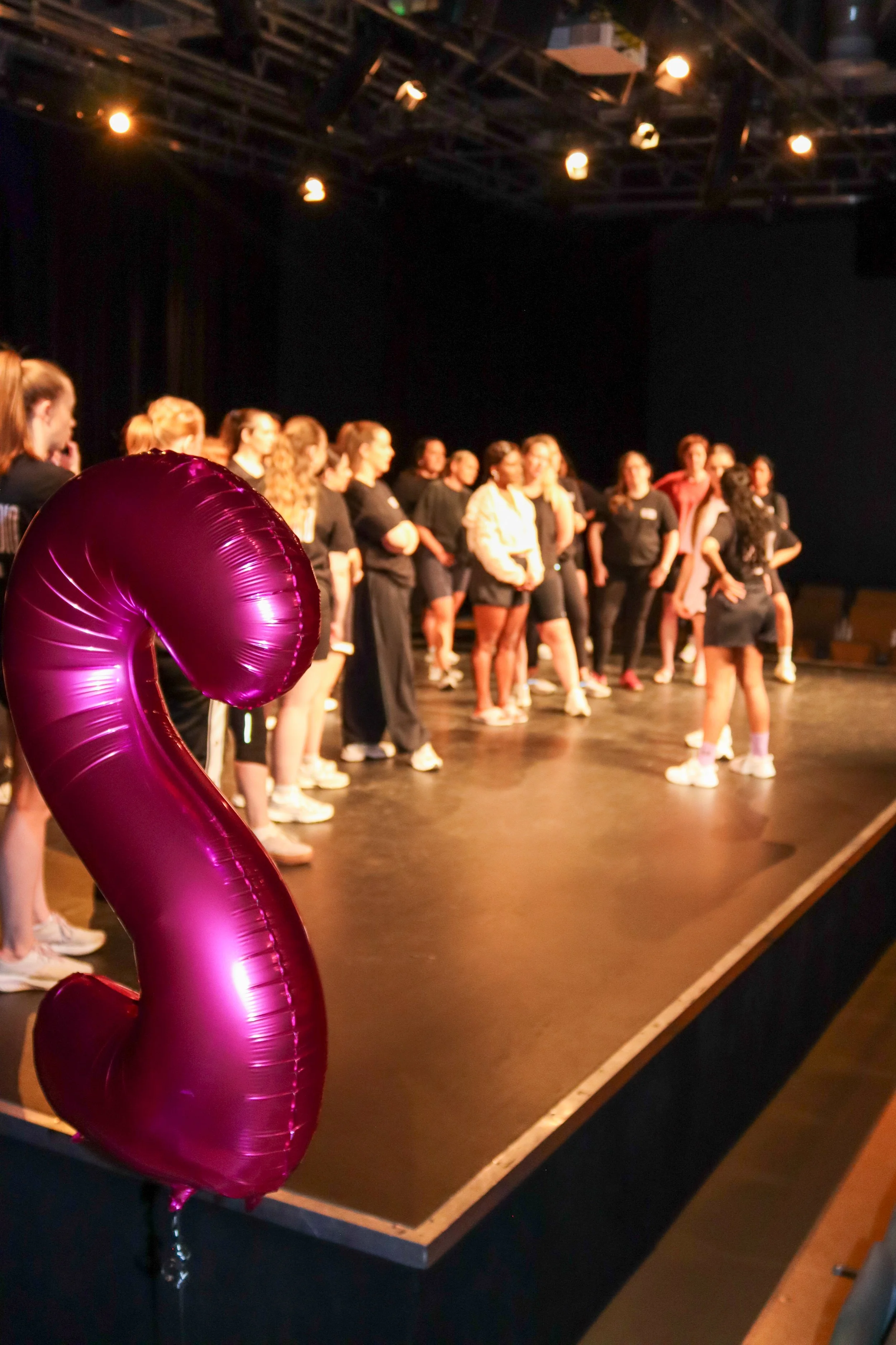 A group of people gathered on a stage with a dark background, with a pink balloon in the foreground.