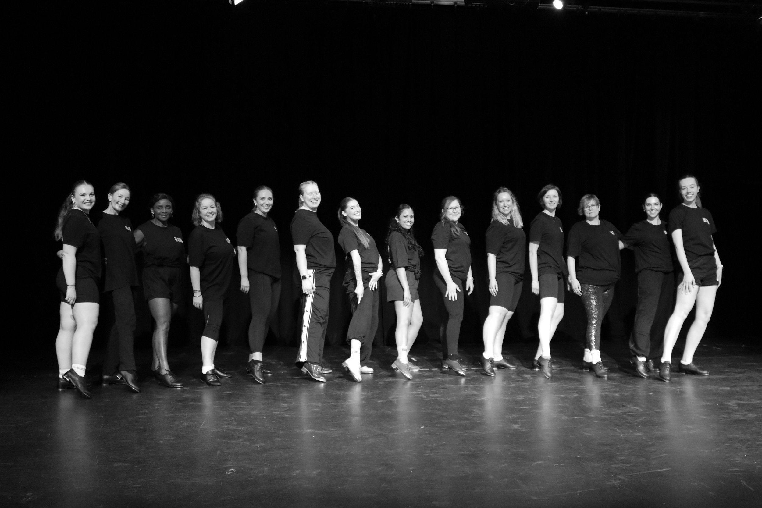 Group of women standing side by side on stage, smiling, wearing black t-shirts and pants, in a theater or performance space.