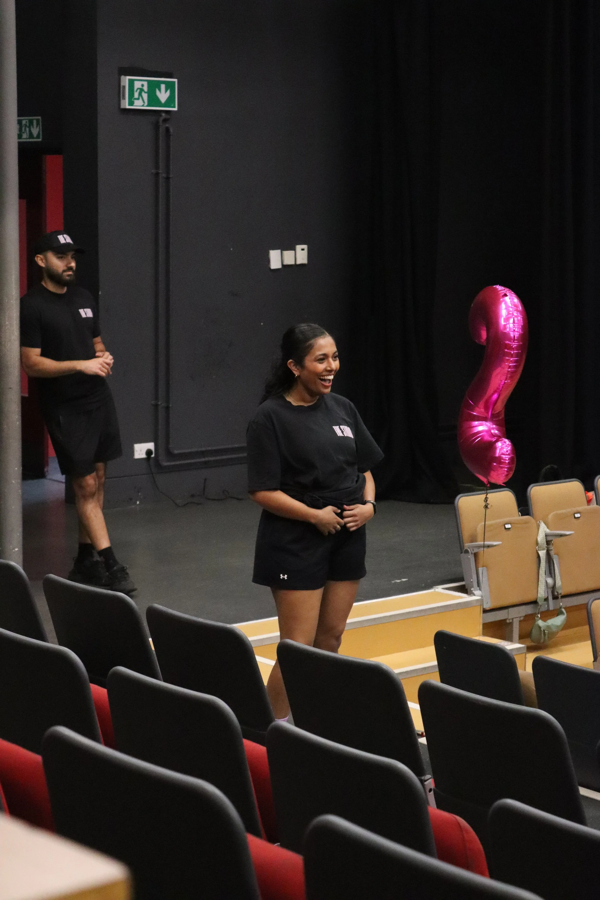 A woman with dark hair in a ponytail, wearing a black shirt and shorts, stands smiling in front of black stage curtains, holding a large pink balloon shaped like the number nine. Two people in black shirts and shorts are in the background, one standi
