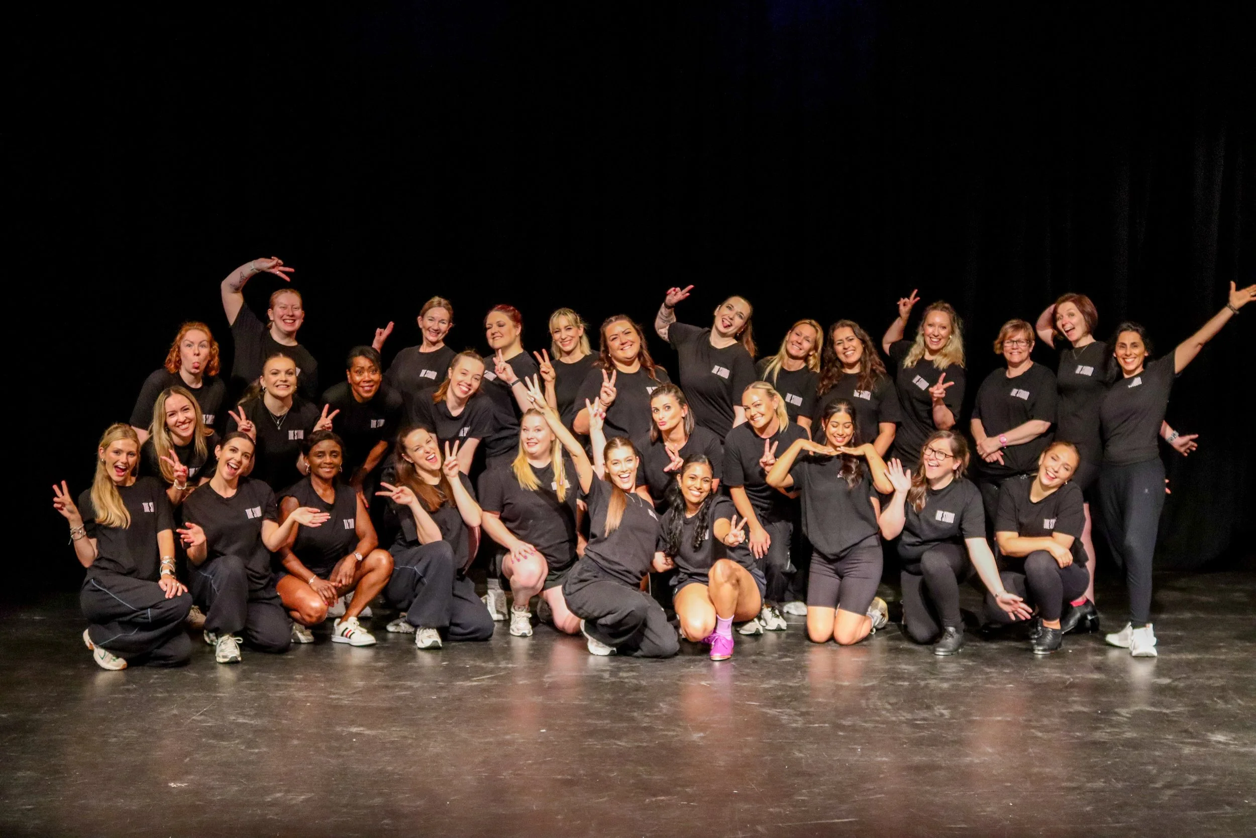 Group of diverse young women posing on stage, smiling and making peace signs, dressed in black t-shirts and casual pants, with a black background.