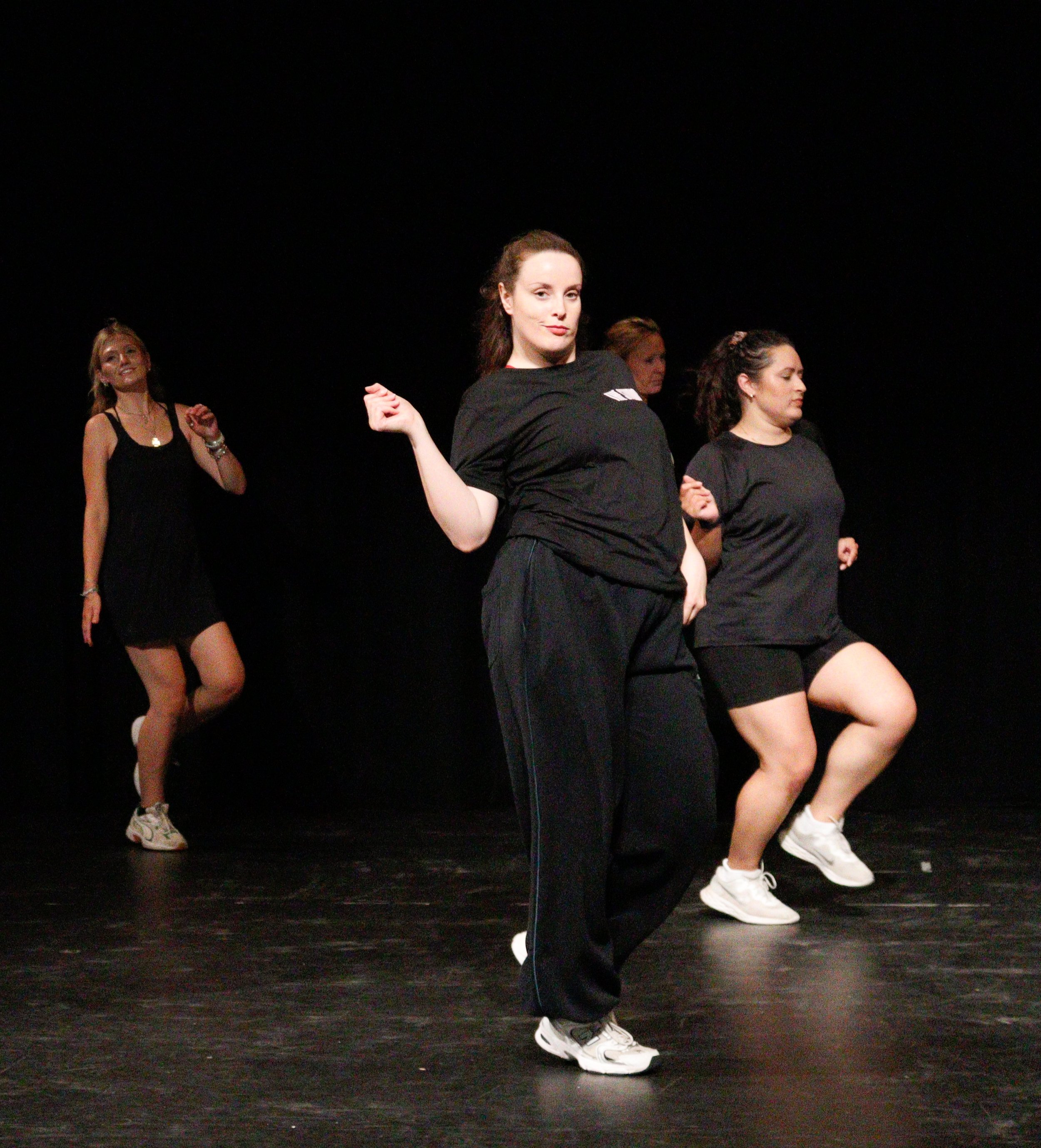 Group of women participating in a dance or exercise class on a dark stage, focusing on a woman in the foreground with a confident expression, wearing black athletic clothing and white sneakers.