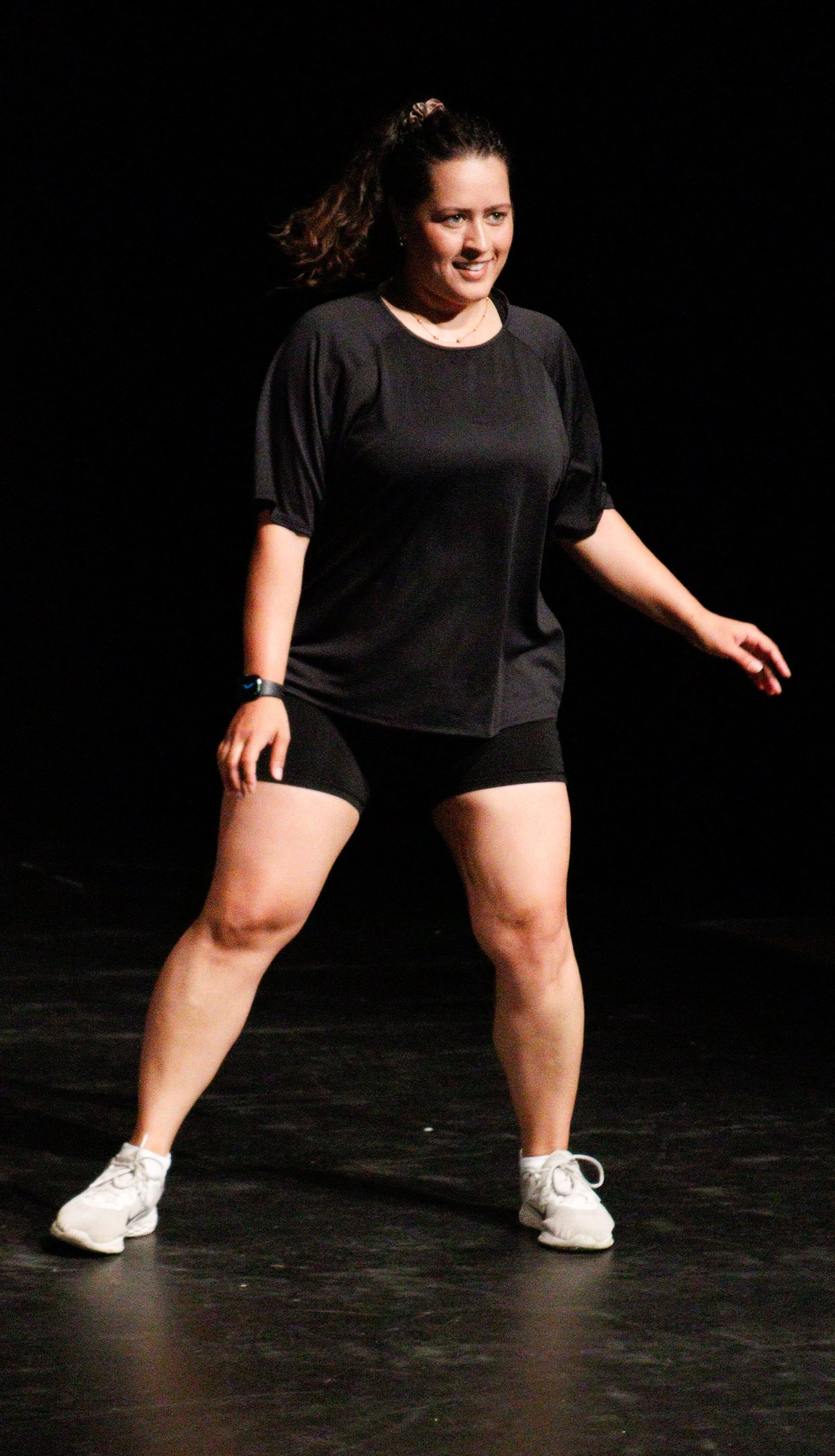 A woman dancing alone on a stage with a black background, wearing a black athletic shirt, black shorts, white shoes, and a fitness tracker on her wrist.